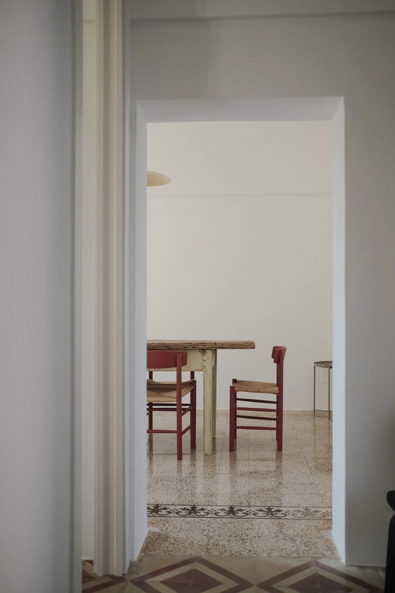 View through a doorway into a room with a wooden table and four chairs with red frames. The room has white walls, a ceiling light, tiled floor, and decorative border tiles at the threshold.