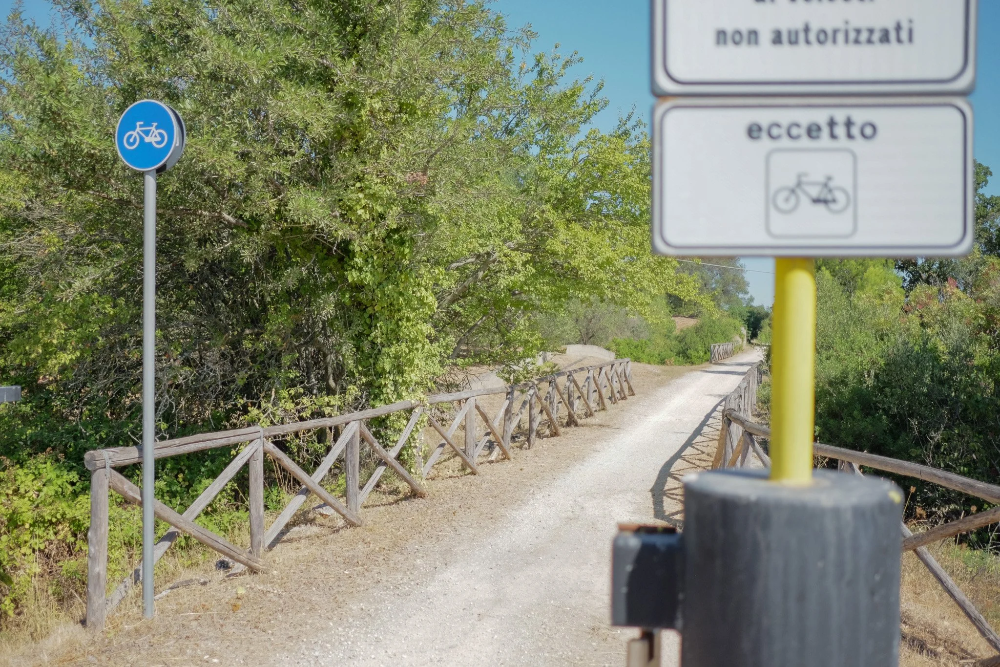 A rural gravel pathway with a wooden fence along each side, surrounded by lush green trees. Two signs are visible: one blue circular sign indicating a designated bicycle lane or path on the left, and a rectangular sign with Italian writing and a bicy
