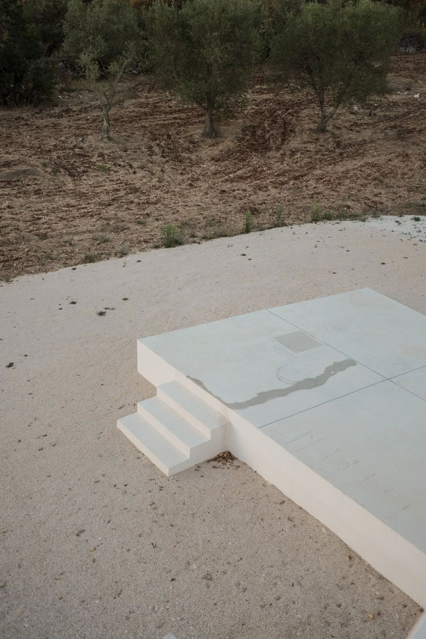 A white outdoor staircase with three steps leading up to a white platform, set on gravel ground, with a dry, dirt hillside and some sparse green bushes and trees in the background.