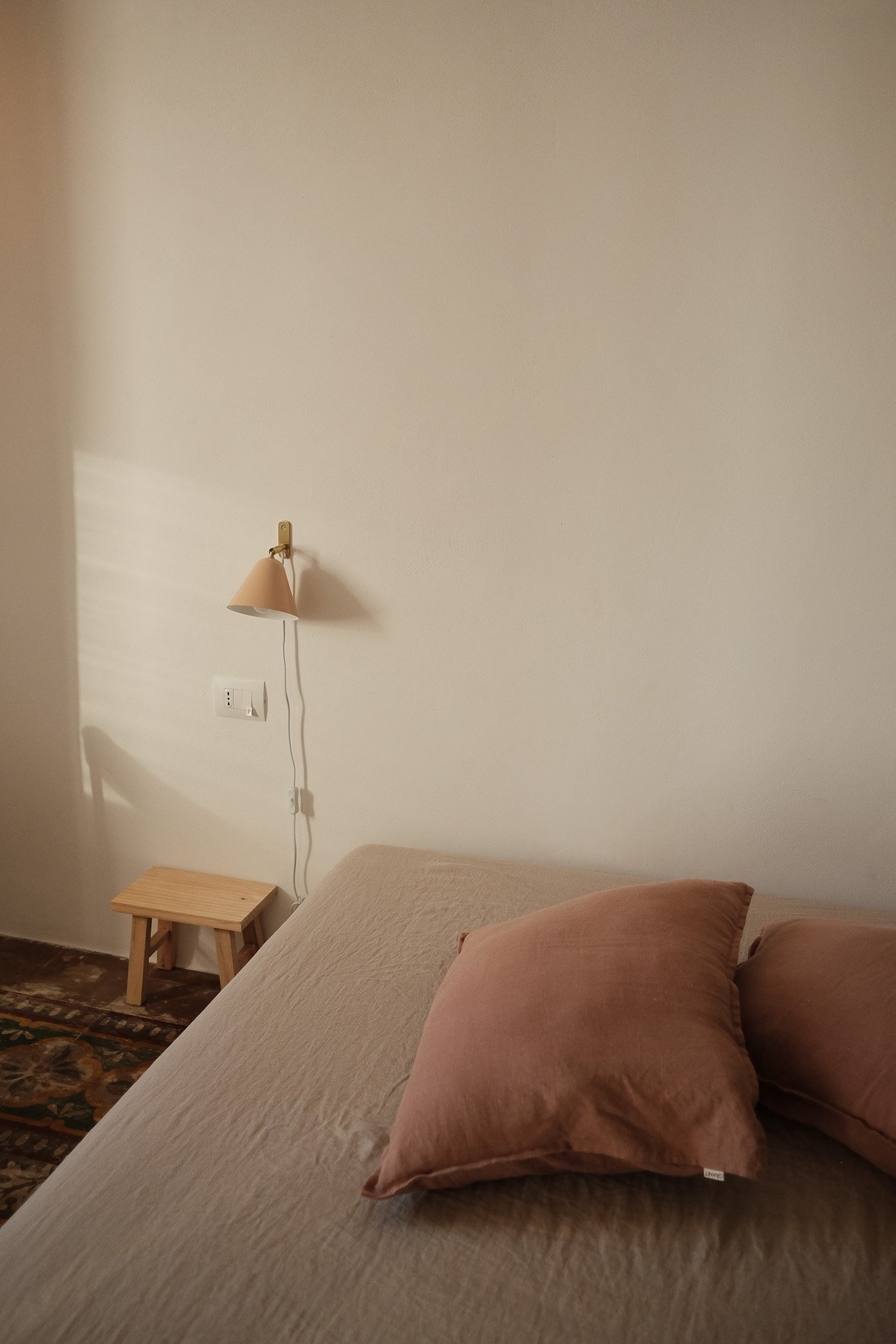 A beige bed with matching pillows in a minimalist bedroom. A wooden nightstand and wall-mounted lamp are beside the bed. The wall behind is plain and painted light, with outlets visible below the lamp.