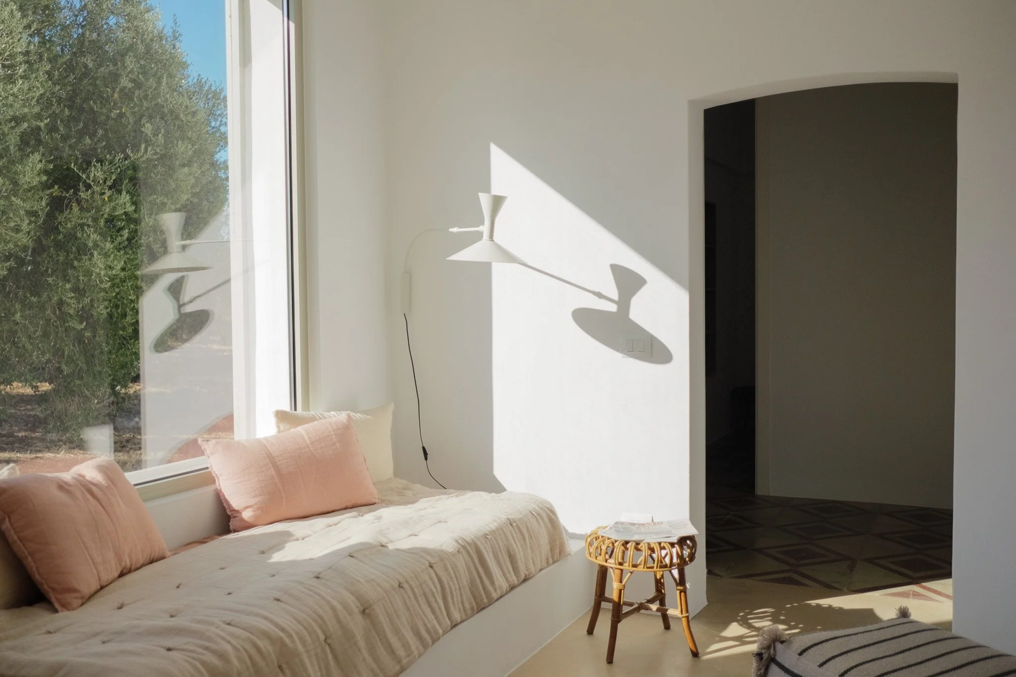 A cozy living room corner with a white bench featuring pink cushions, a large window letting in sunlight, a rattan side table, and a modern white floor lamp casting a shadow on the white wall.