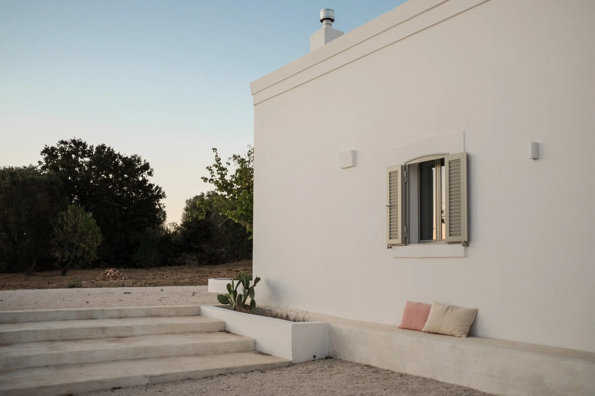 A white Mediterranean-style house with a small window with shutters, a chimney, and outdoor seating area with cushions, set against trees and blue sky at sunset.
