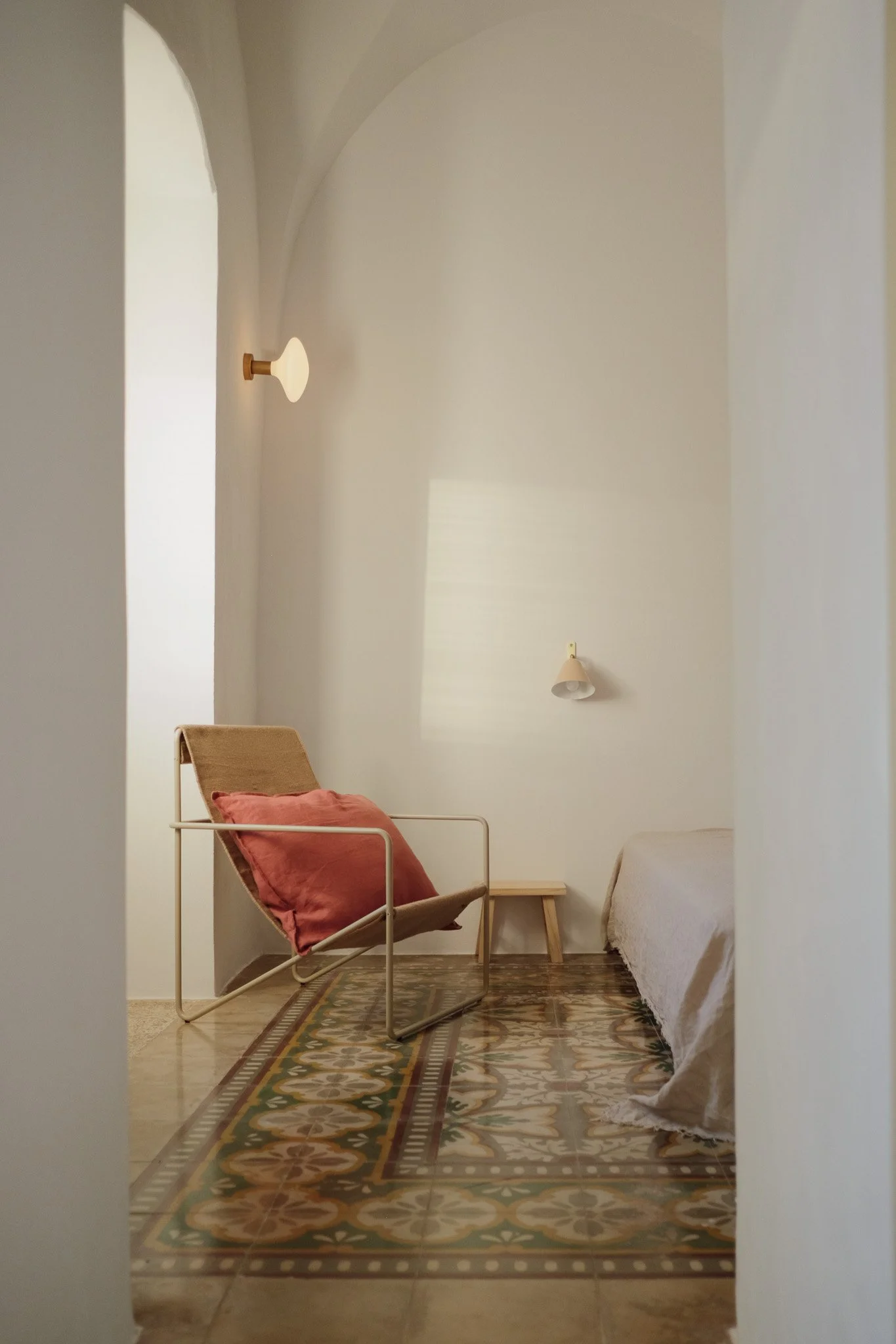A cozy bedroom corner with a beige armchair that has a reddish-brown pillow, a small wooden side table, a bed with white bedcover, and wall-mounted lights, all on a patterned tile floor with a decorative rug.