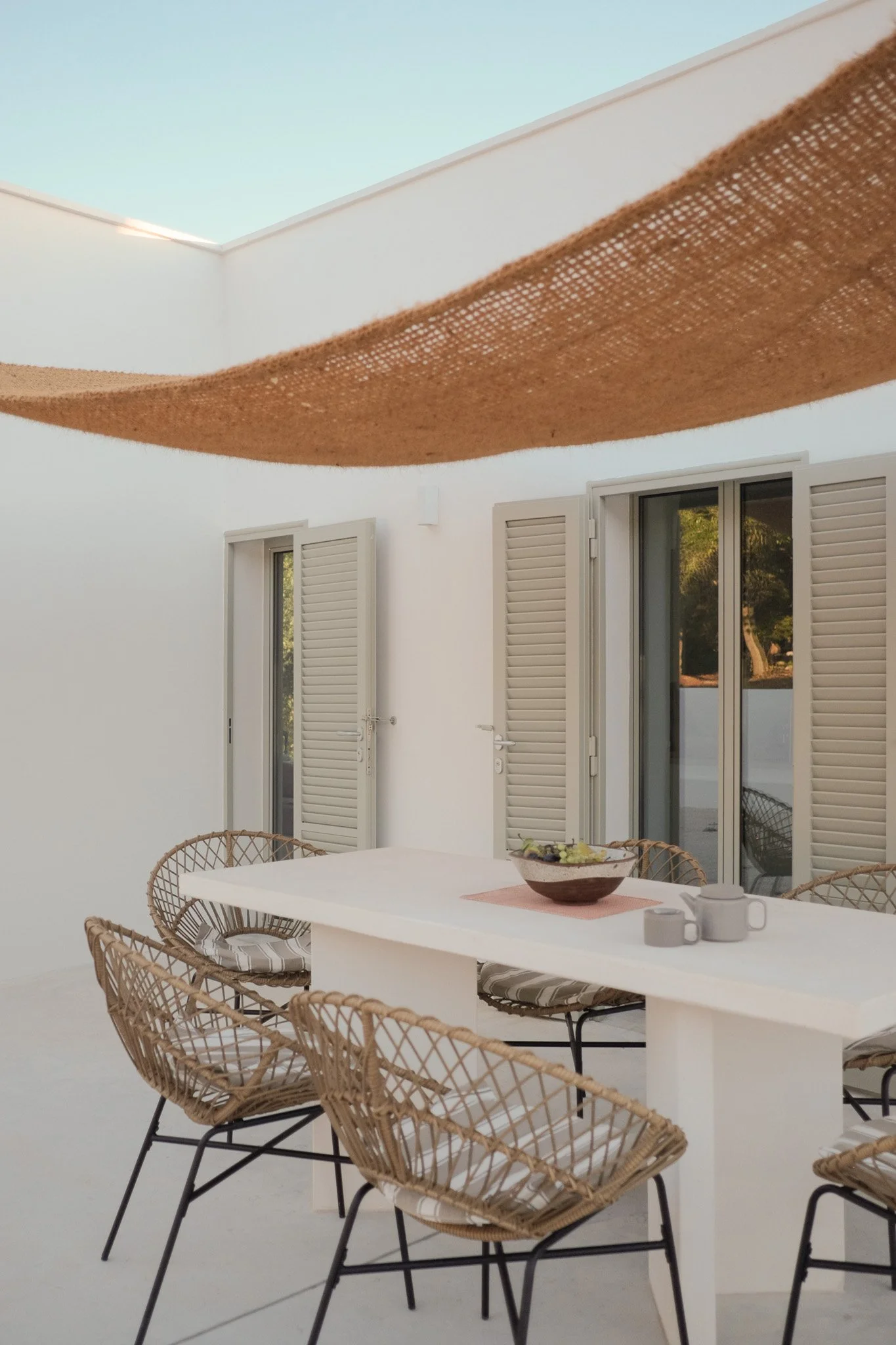 A white dining table with a bowl of food, cups, and utensils, surrounded by woven chairs with cushions, set on a patio or balcony with white walls, windows, and shutters, and a brown fabric sunshade overhead.