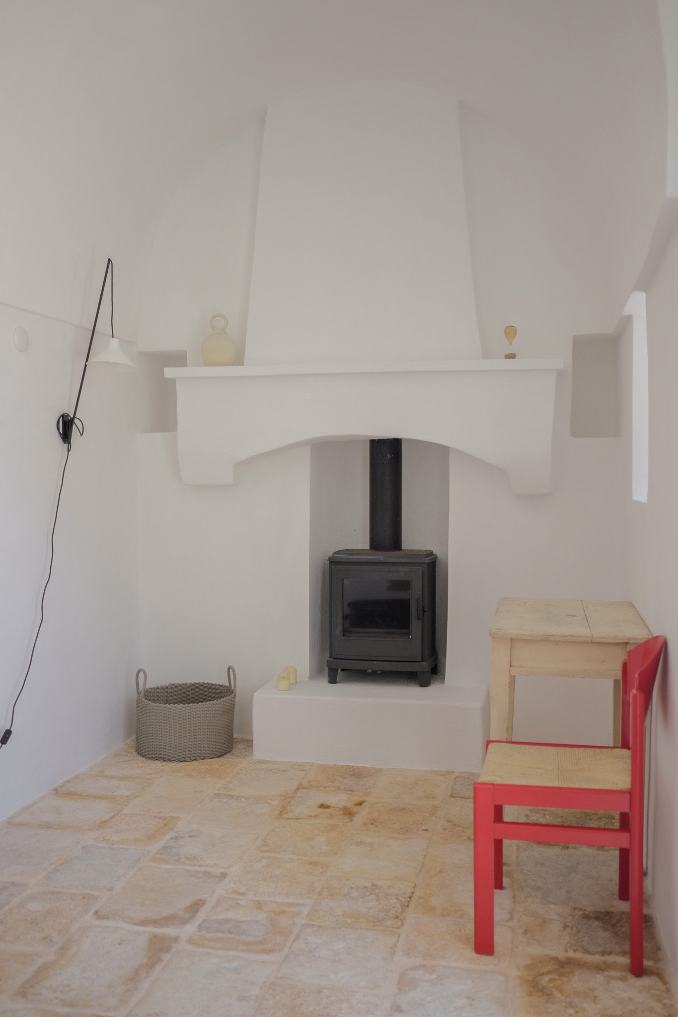A cozy room with a wood stove fireplace, a gray woven basket, a small candle, a red chair, and a wooden table on beige tiled floor, with a white wall and decorative vases on a step above the fireplace.