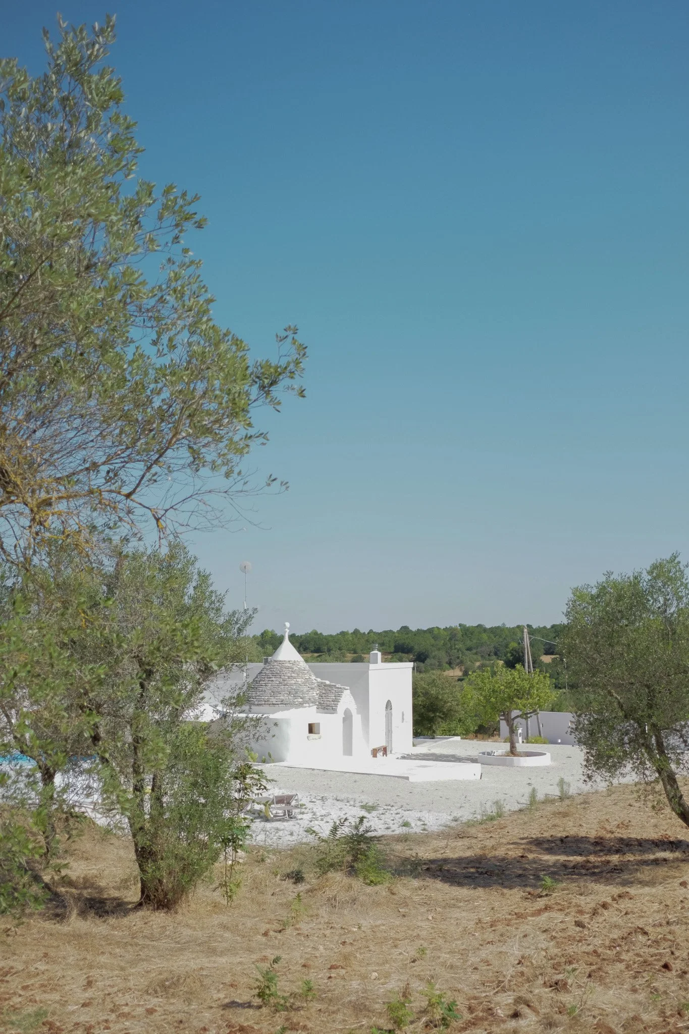 A white building with a cone-shaped roof and small windows, surrounded by trees and open space under a blue sky.