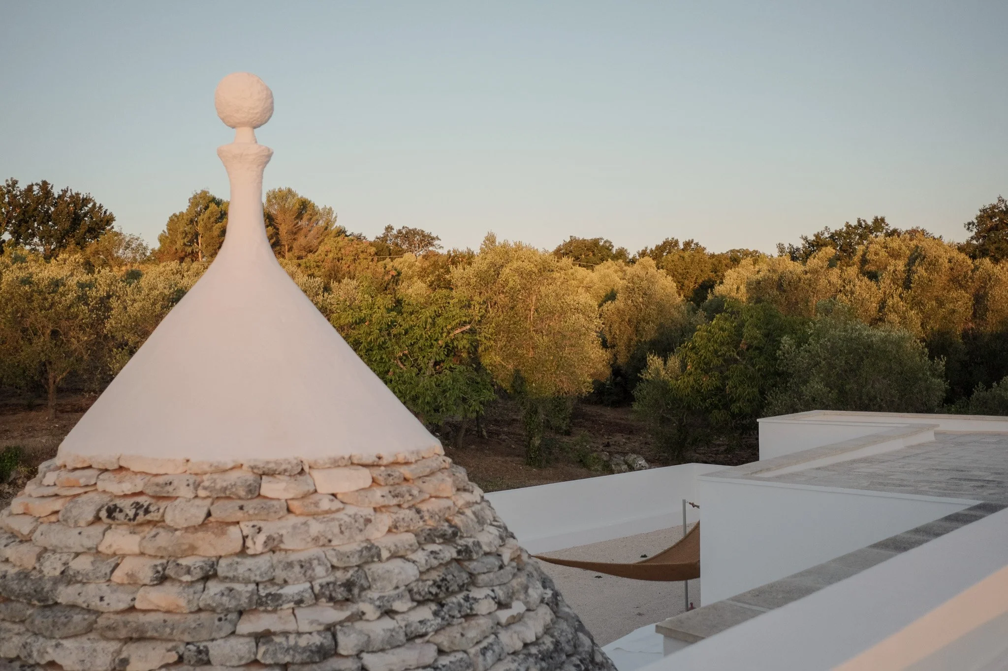 A white dome-shaped structure with a stone base and a rounded top, set against a background of trees and a pale sky.
