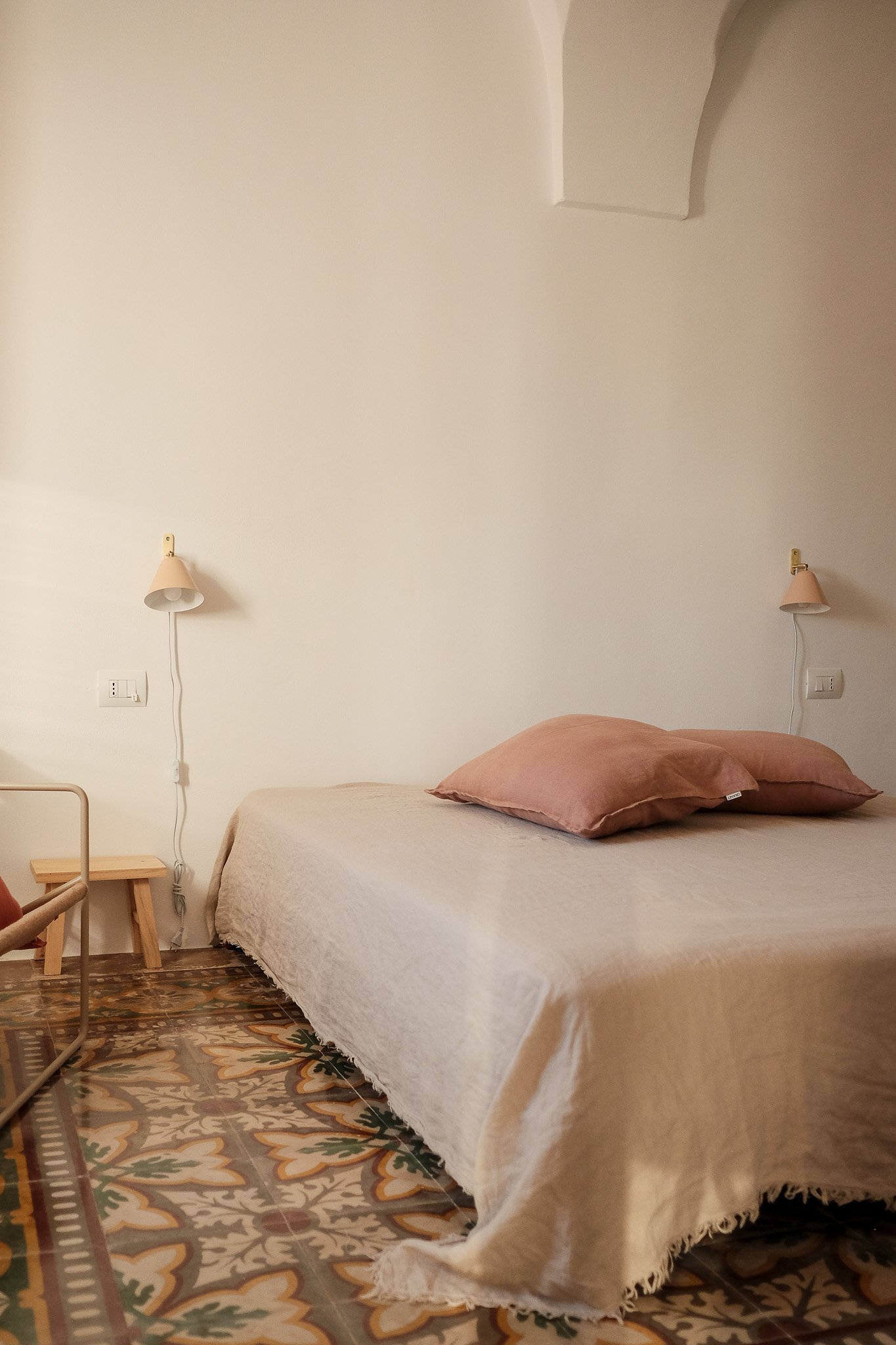 A bedroom with a bed covered in a cream-colored textured blanket, two pink pillows, two wall-mounted lamps with pink shades, a small wooden side table, a patterned tile floor, and a plain white wall with a small recessed section