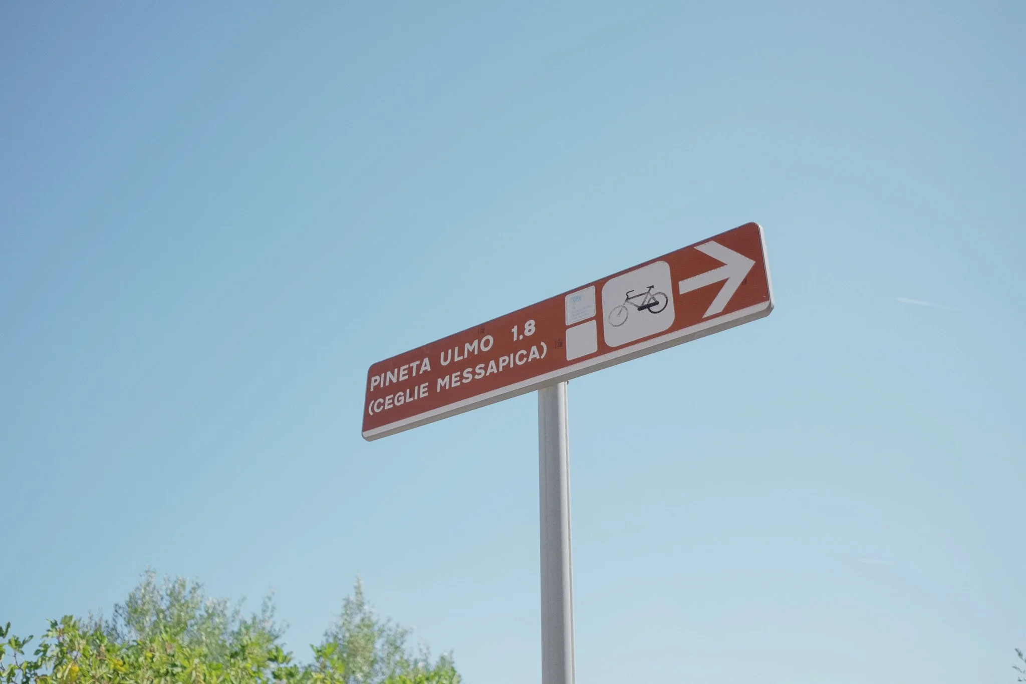 A red and white sign with a bicycle symbol and an arrow pointing right, indicating a bicycle path or lane, with text in Italian mentioning Pineta Ulmo and Ceglie Messapica, against a clear blue sky with some trees below.