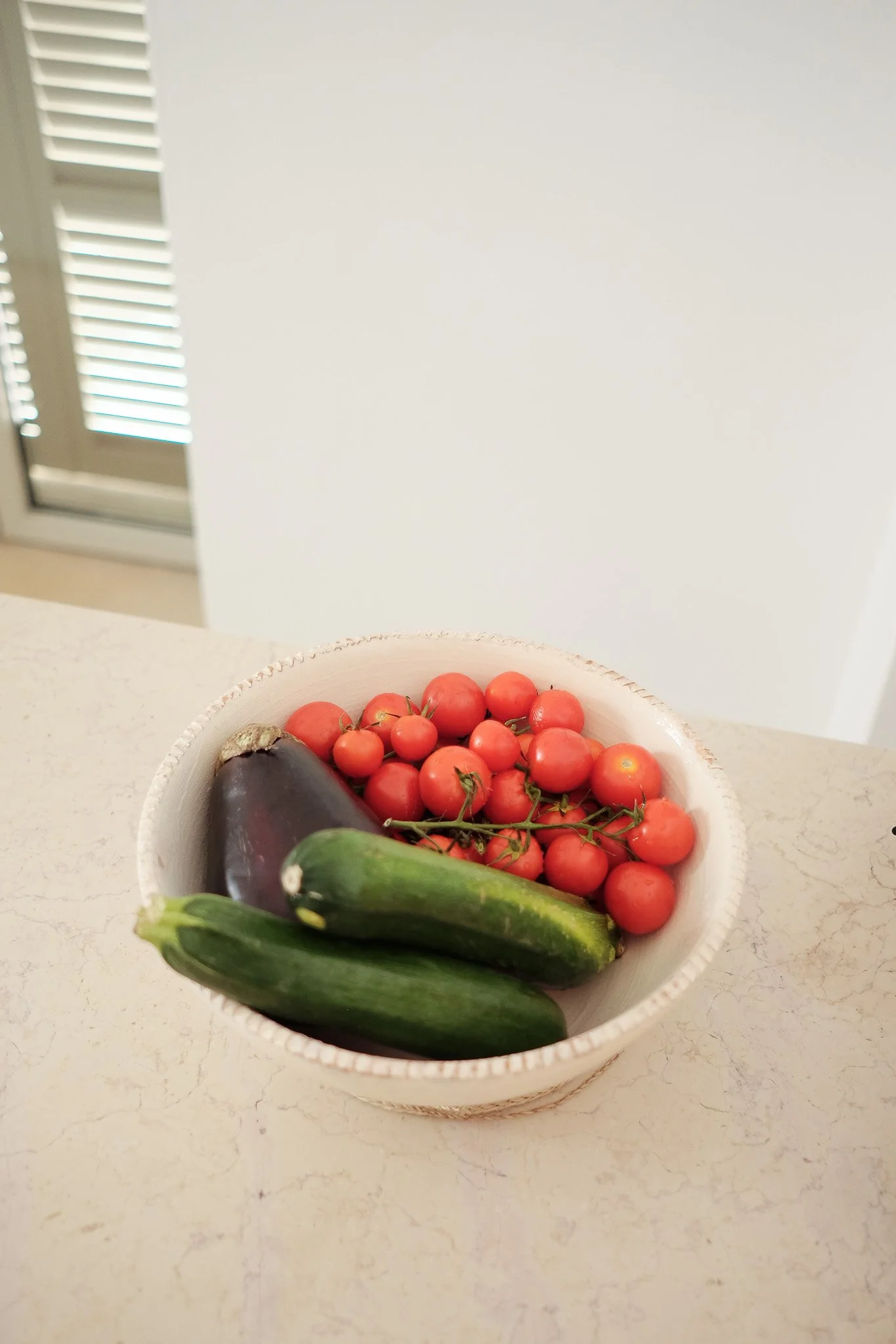 A bowl of fresh vegetables including cherry tomatoes, eggplant, and zucchinis, placed on a kitchen countertop with a white background.