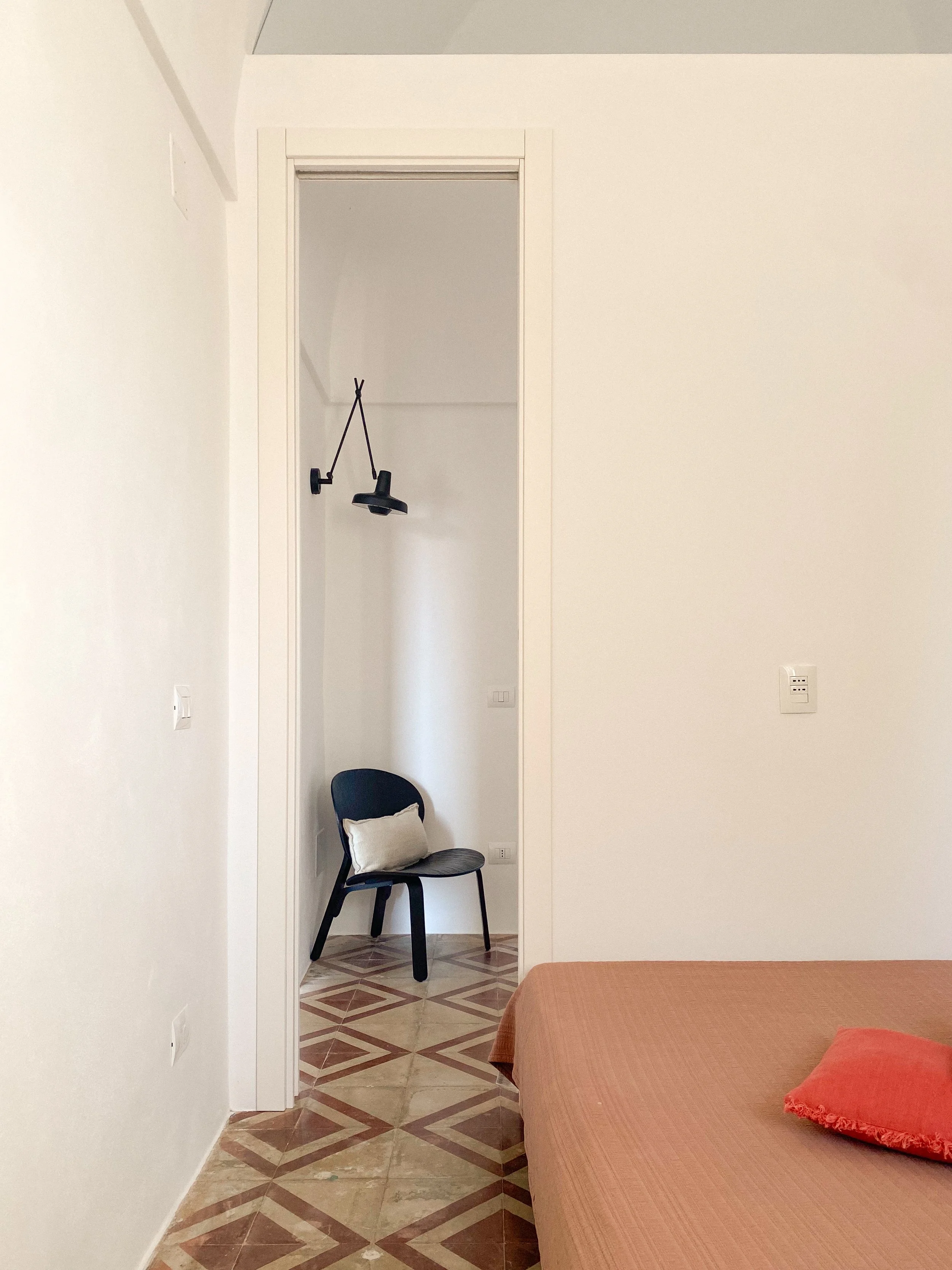 A bedroom with a bed, chair with a cushion, and a doorway with a lamp hanging from the top of the doorway. The walls are white and the floor has a patterned tile design.