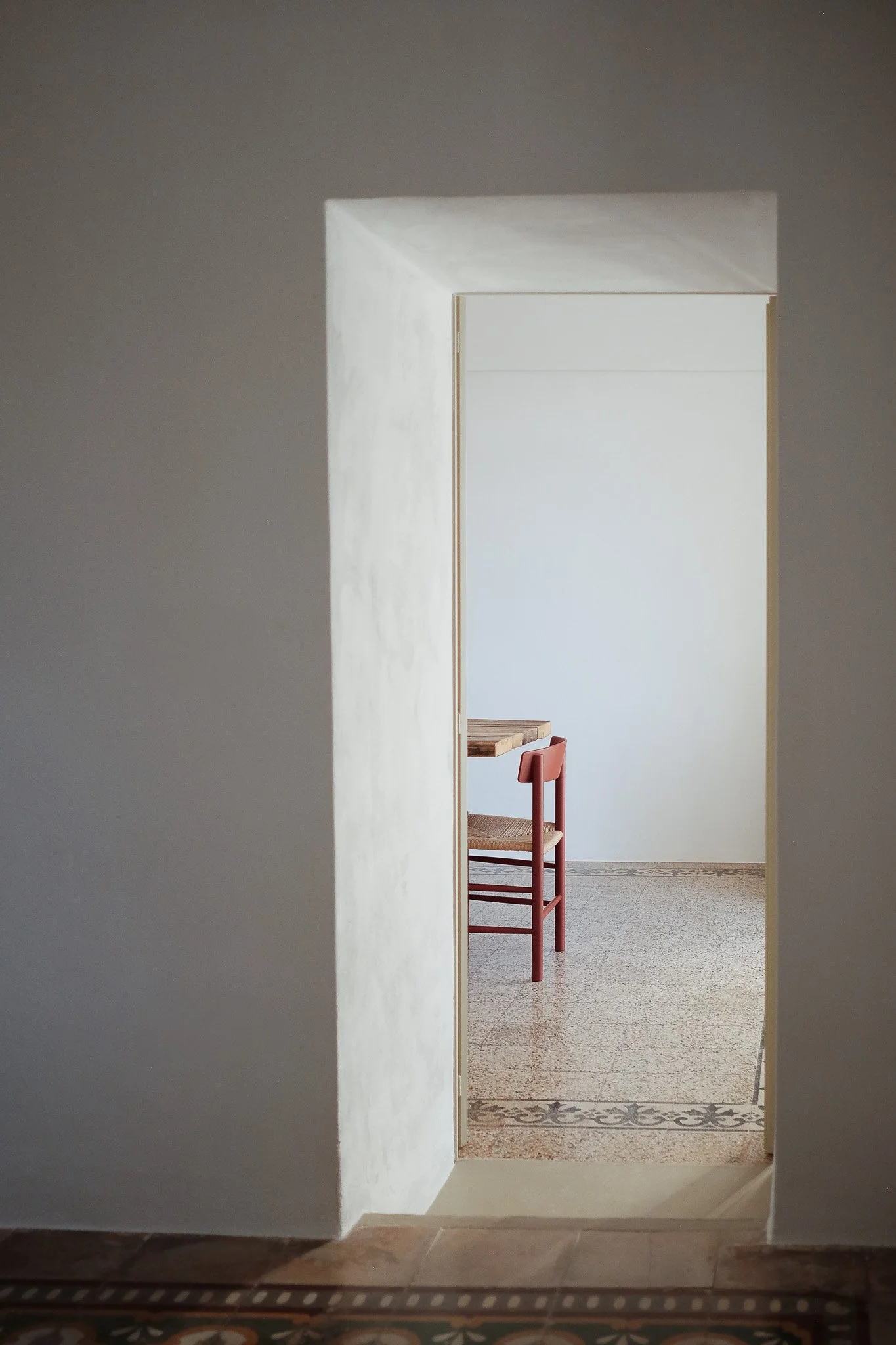 Image of a view through a doorway into a room with a wooden table and a red chair, with a white wall in the background and patterned floor tiles at the entrance.