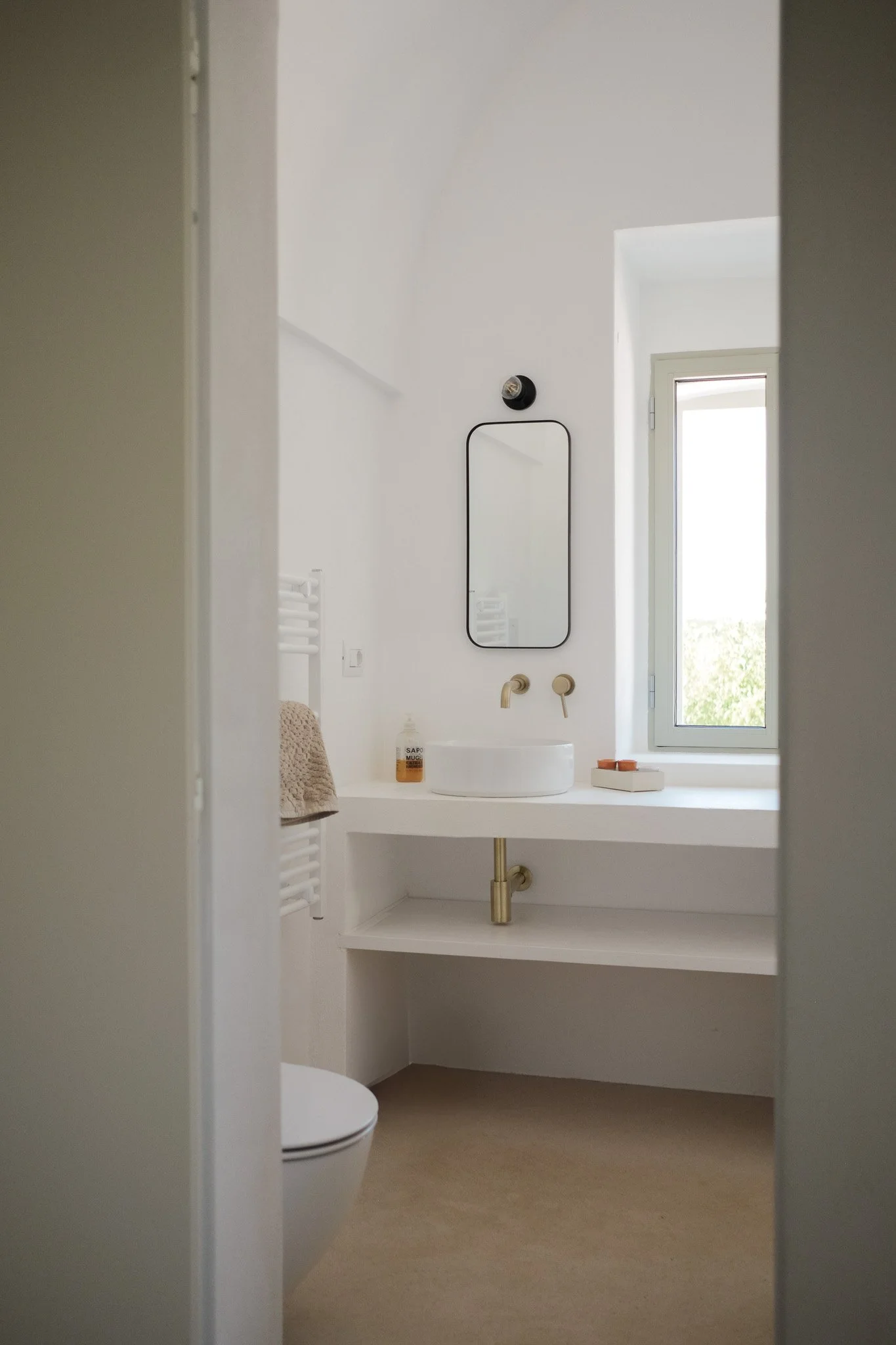 A modern, minimalist bathroom with white walls and a large window. A round white sink sits on a countertop with a single faucet and soap dispenser. A mirror hangs above the sink. There is a towel on a rack to the left and a toilet in the foreground. 
