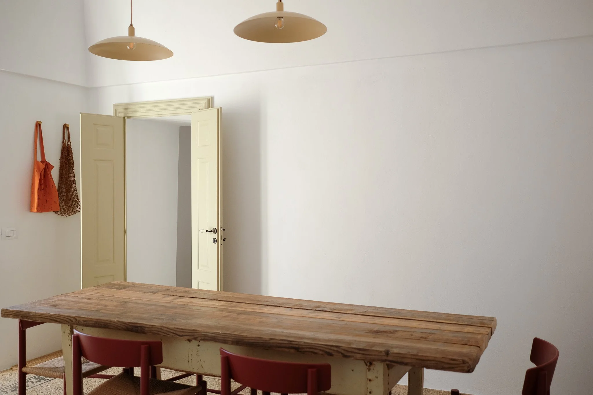 A dining area with a wooden table and red chairs, beige walls, a doorway with open double doors, two pendant lights hanging from the ceiling, and wall hooks with orange and brown bags hanging on them.