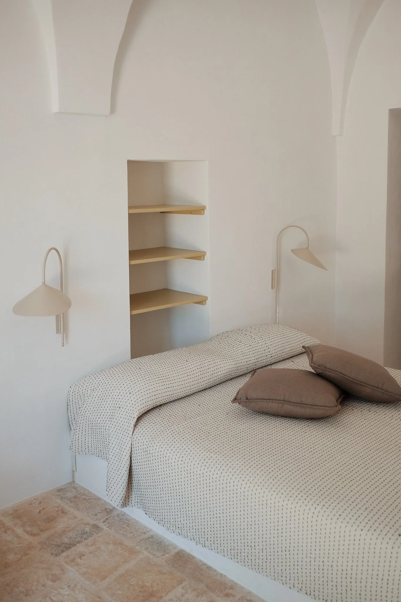 A bedroom with a bed covered with a patterned white and black blanket and two brown pillows. White wall-mounted lamps on either side of the bed and internal shelves built into the wall behind it. A small window-like arch is visible near the ceiling.