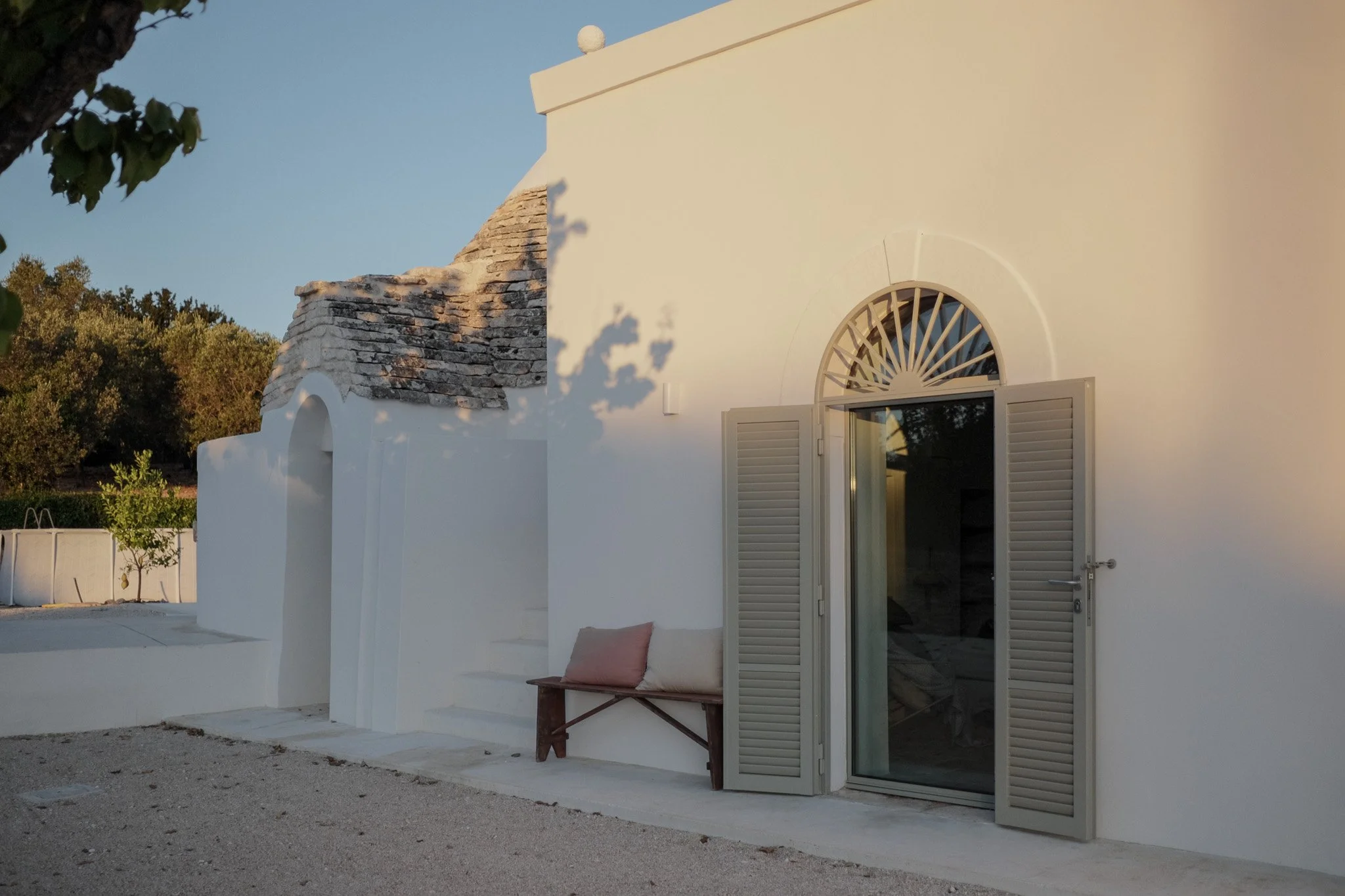 White building with a rounded window and open shutters, a small wooden bench with a pink cushion, and a rustic stone chimney, set against a clear sky in a Mediterranean style setting.