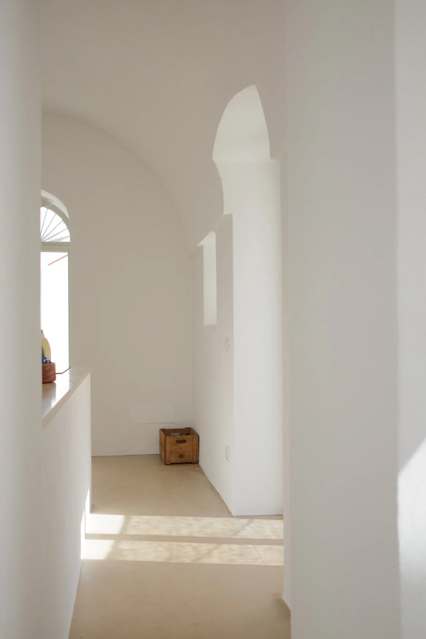 A narrow hallway with white walls and light-colored flooring, sunlight casting shadows on the floor, a small wooden box at the end, and a window on the left side of the hallway.
