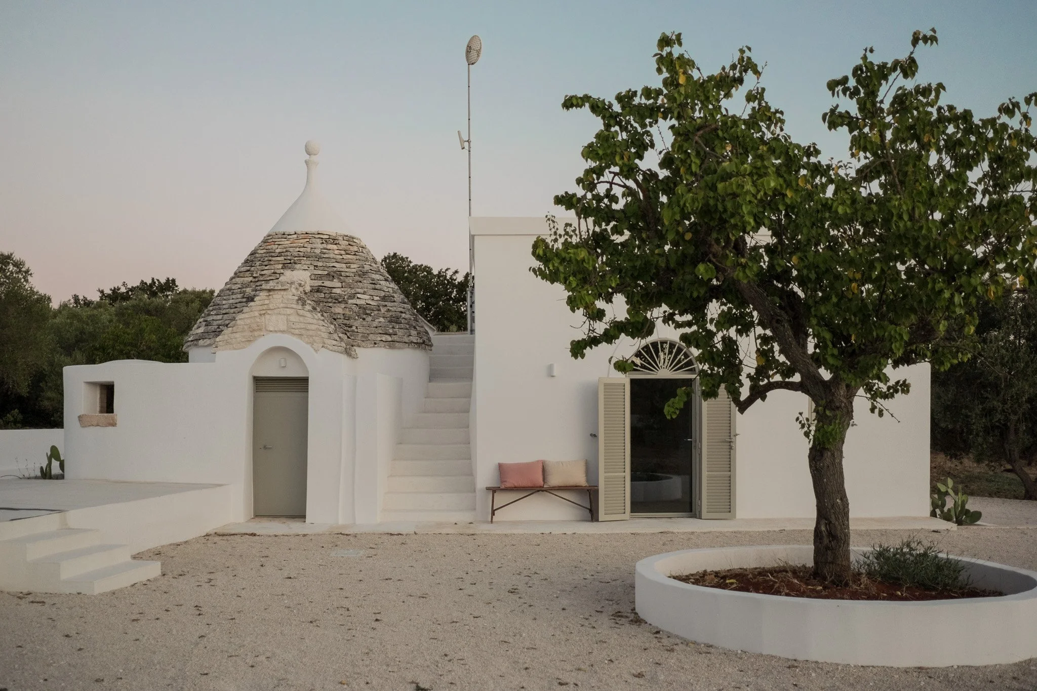 A white building with a conical roof made of stone tiles, a gray door, a window with open gray shutters, and a small bench with two cushions in front. There is a large tree in a circular bed to the right and a white gravel ground with a staircase lea