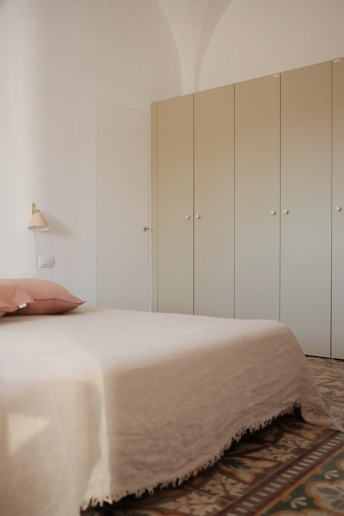 A bedroom with a bed covered in beige bedspread and pillows, a white wardrobe with multiple doors, a small wall lamp, and patterned area rug visible at the foot of the bed.