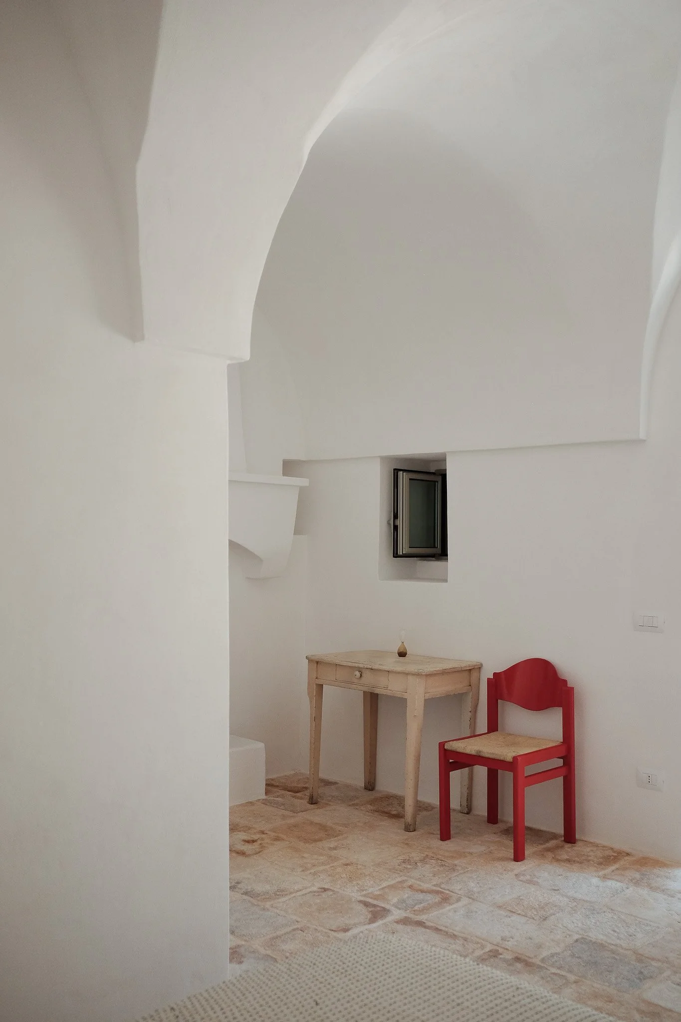 A corner of a room with white walls and ceiling. There is a small window, a red chair, a distressed wooden table, and a decorative vase on the table. The floor is covered with light-colored tiles and a small beige rug.