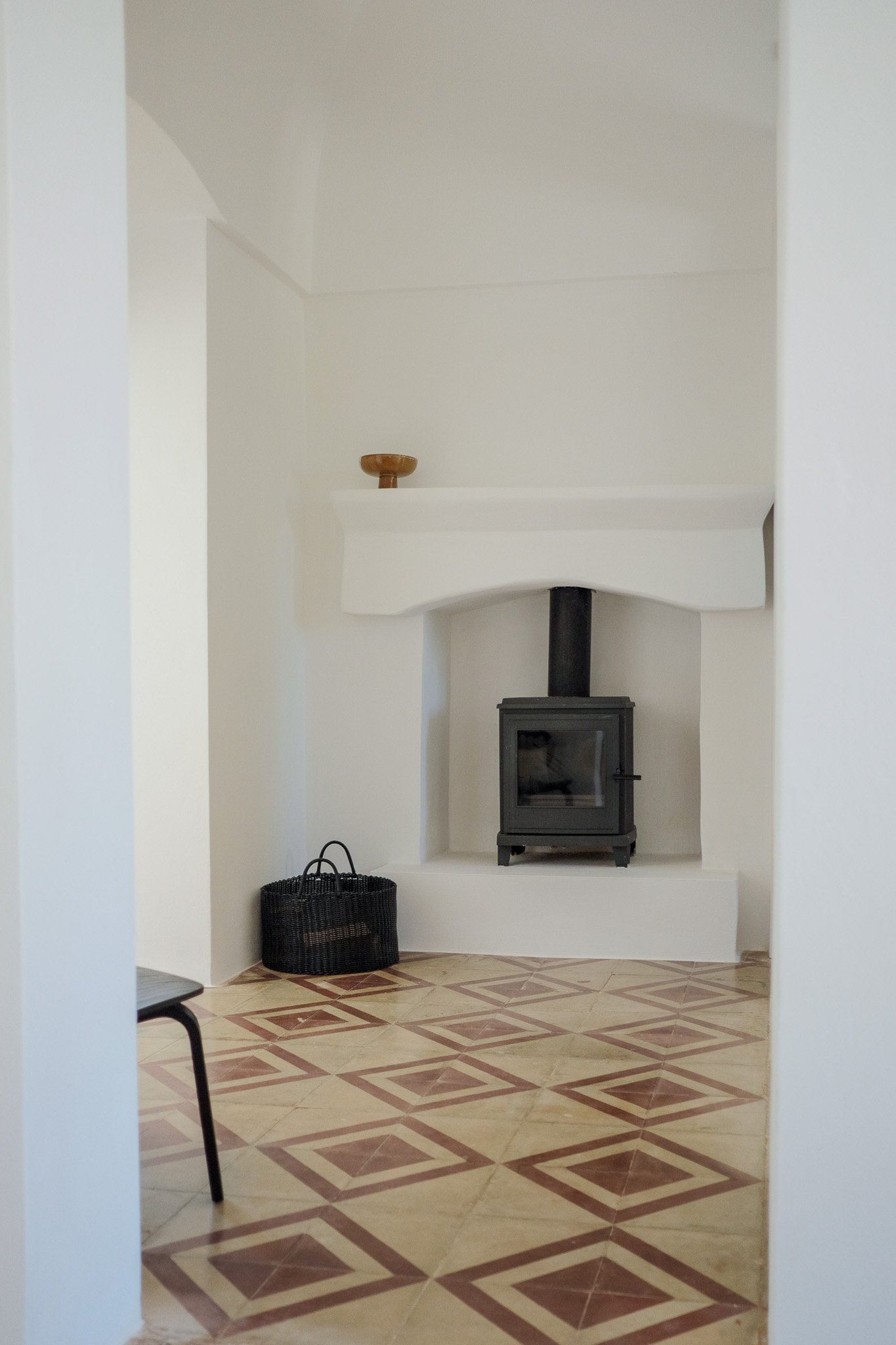 A small black wood stove in a corner of a room with white walls and beige patterned tile floor, a black basket on the floor to the left of the stove, and a small ledge with a wooden bowl on top of it above the stove.