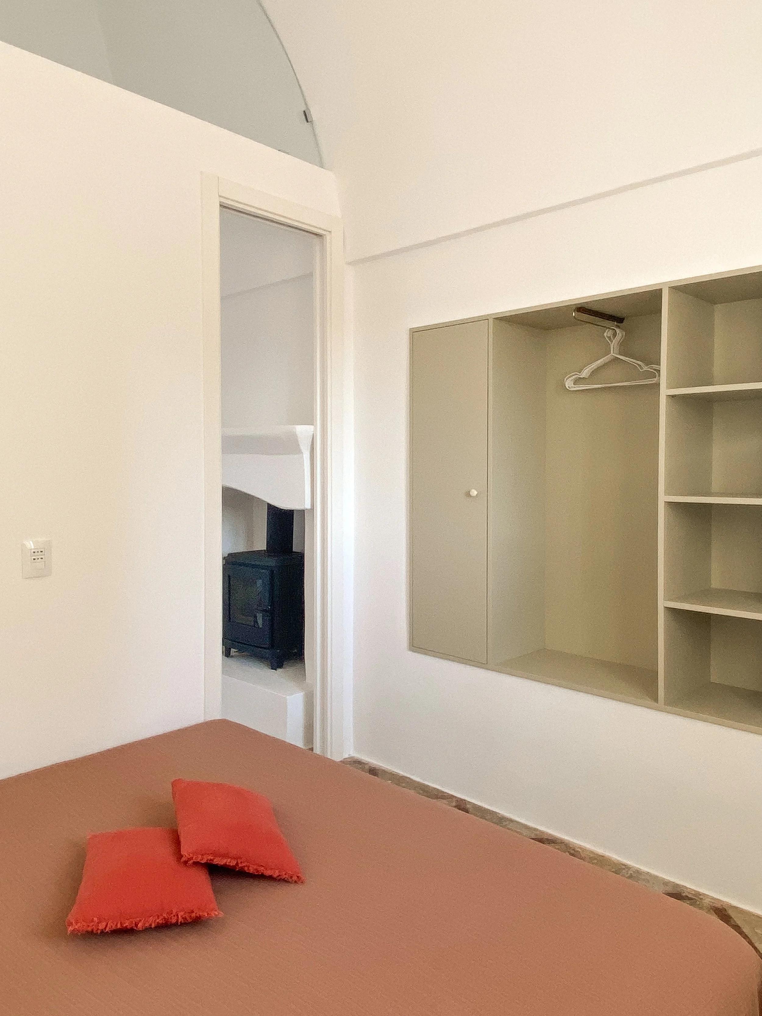 A bedroom with a brown bedspread and two orange pillows, a beige wall with a built-in closet with one open door and some empty shelves, and an oak-colored baseboard. There is a doorway leading to another room with a black wood stove visible.