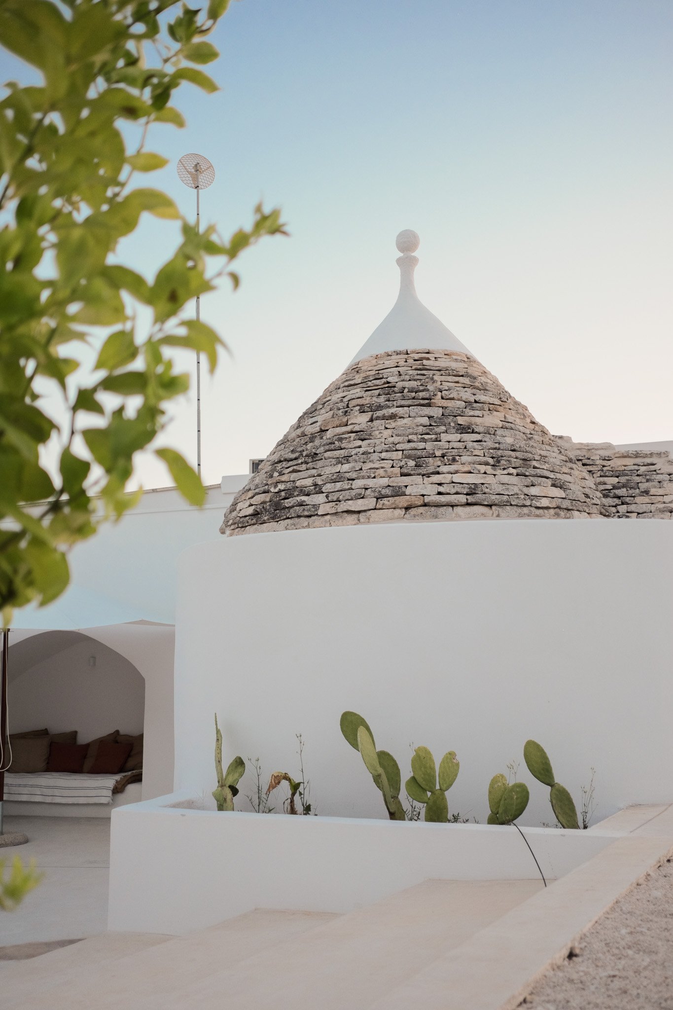 White building with a rounded roof made of stone, topped with a decorative white finial, and a cactus garden in front. In the background, part of a terrace with a bed and pillows, and a blue sky.