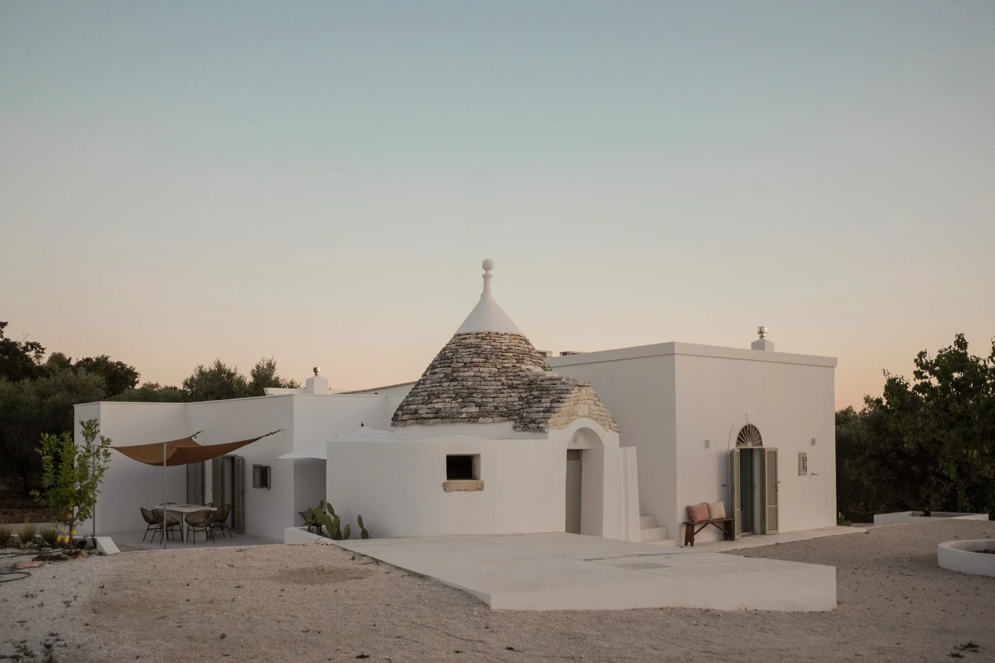 A white building with a cone-shaped roof made of stone blocks and a small round ornament at the top, set against a clear sky during sunset. There is outdoor seating with chairs and a table, and trees surrounding the building.