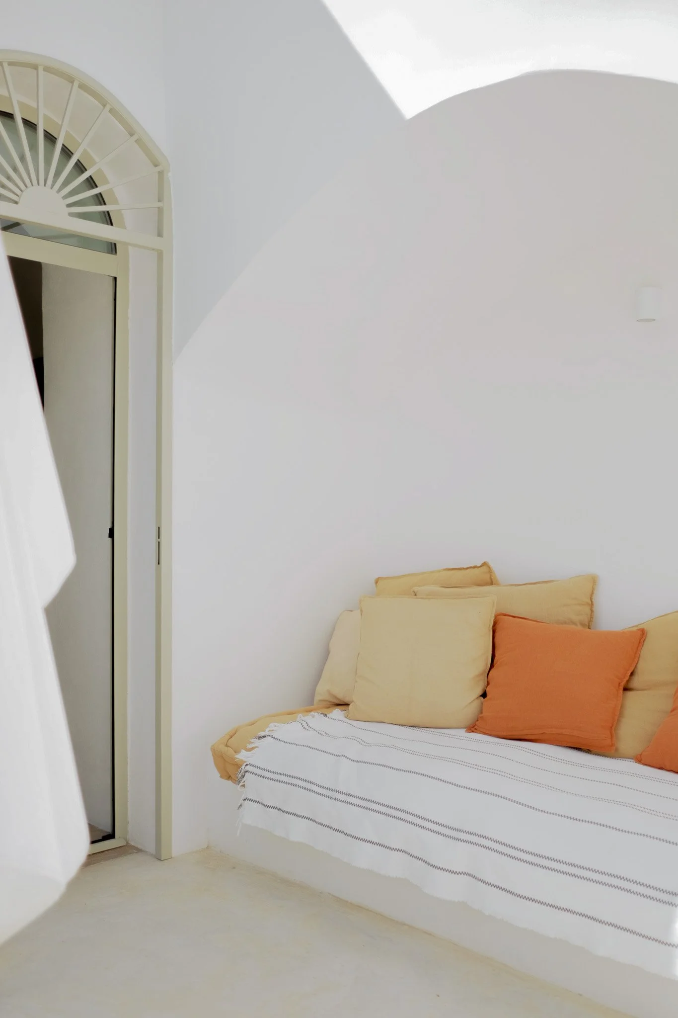 A bedroom with a bed covered with a striped white and black bedspread, beige and orange pillows, and a plain white wall in the background. Part of a window or doorway with a cream-colored frame is visible on the left side.