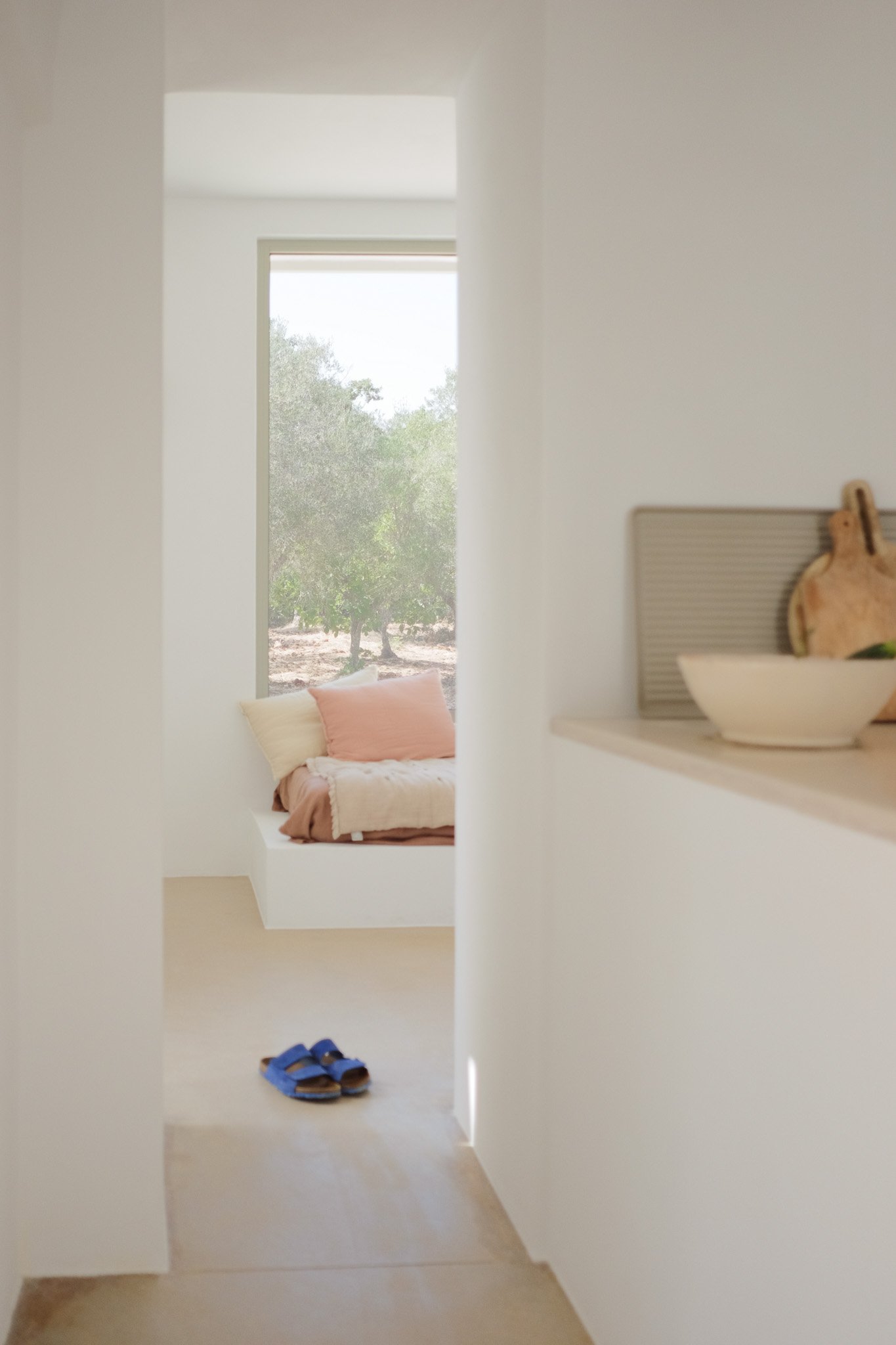 View of a cozy bedroom with a window overlooking greenery, a bed with pink and off-white pillows, and a pair of blue sandals on the floor near the entrance.