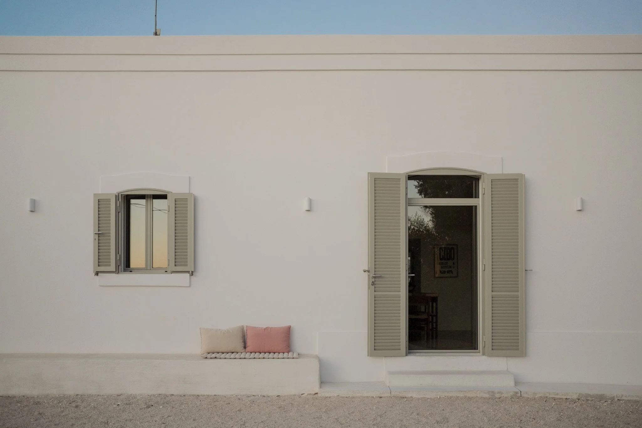 White building with two windows and one door, all equipped with light-colored shutters, and a pink and beige cushion on the ground in front.
