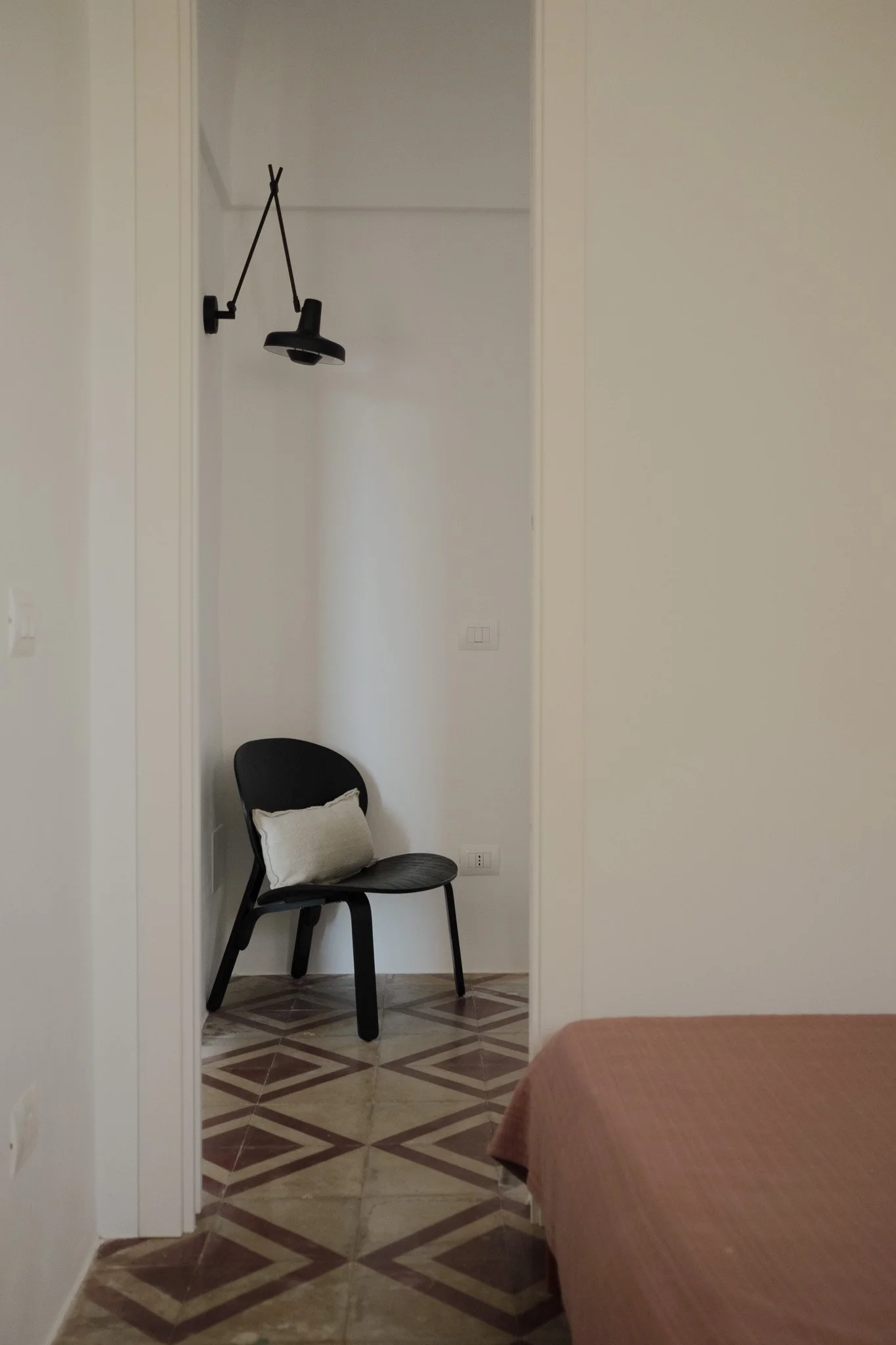 A small black chair with a light gray pillow resting on the seat, positioned against a white wall in a room with tiled floor. A black wall-mounted lamp is hanging above the chair. Part of a bed with a brown bedspread is visible in the lower right cor