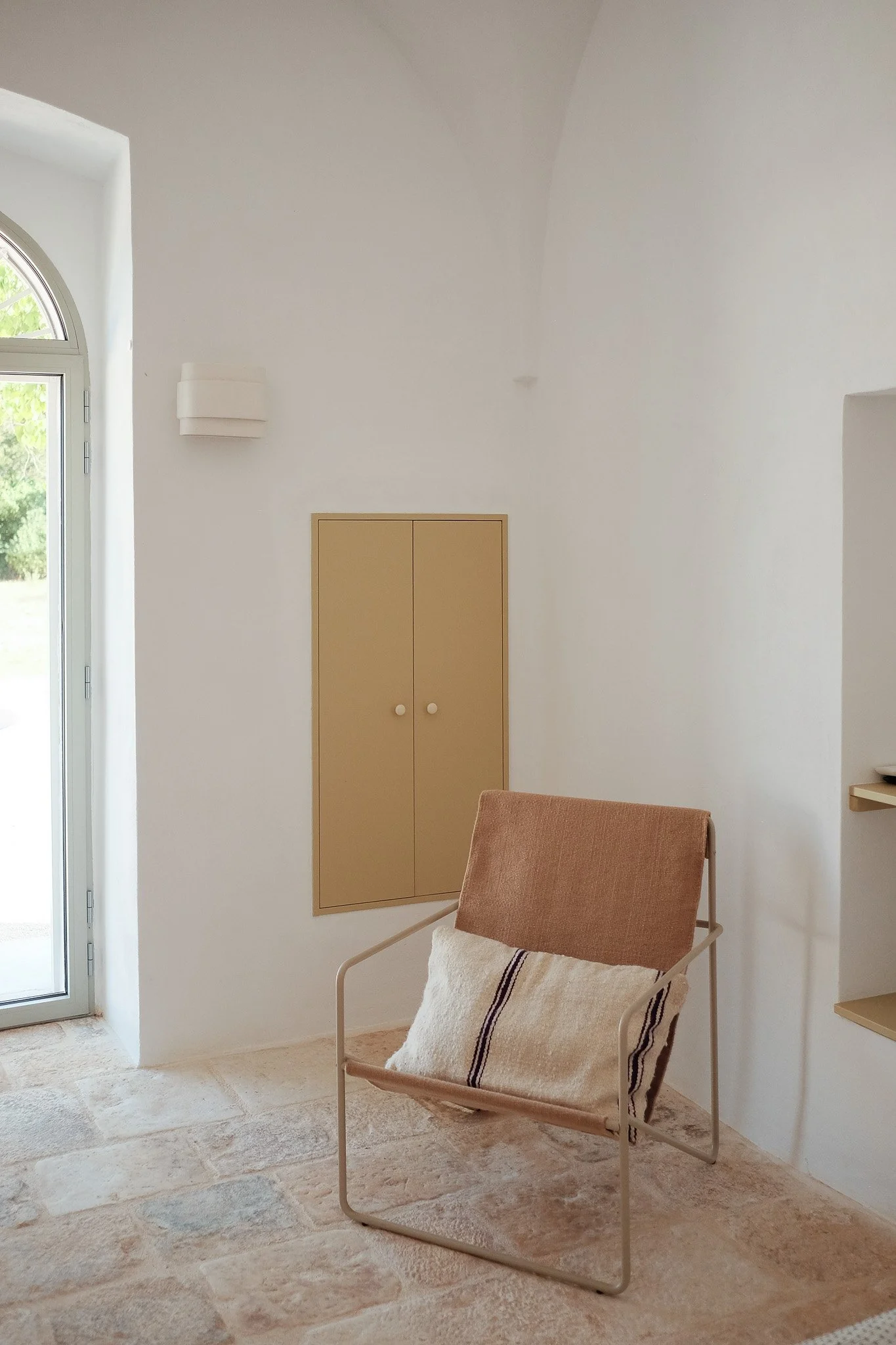 A cozy corner with a beige chair, a white pillow with a black stripe, a wall-mounted light, a small built-in cabinet, and a glass door with a view of greenery outside