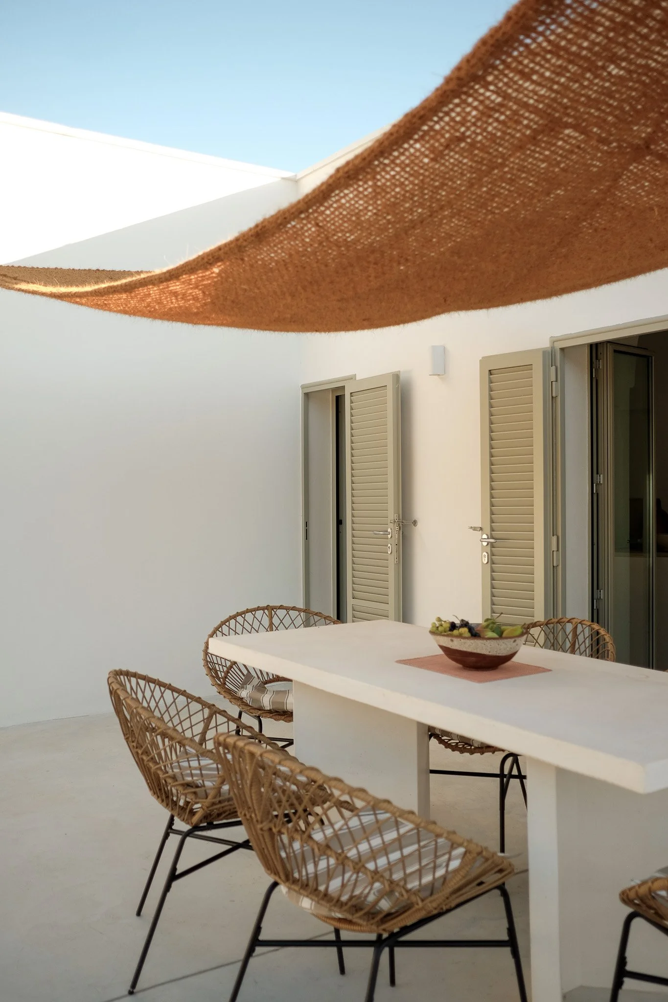 A white outdoor patio with a white table, tan wicker chairs, and an open door leading to an indoor space, under a large tan fabric shade in a sunny setting.