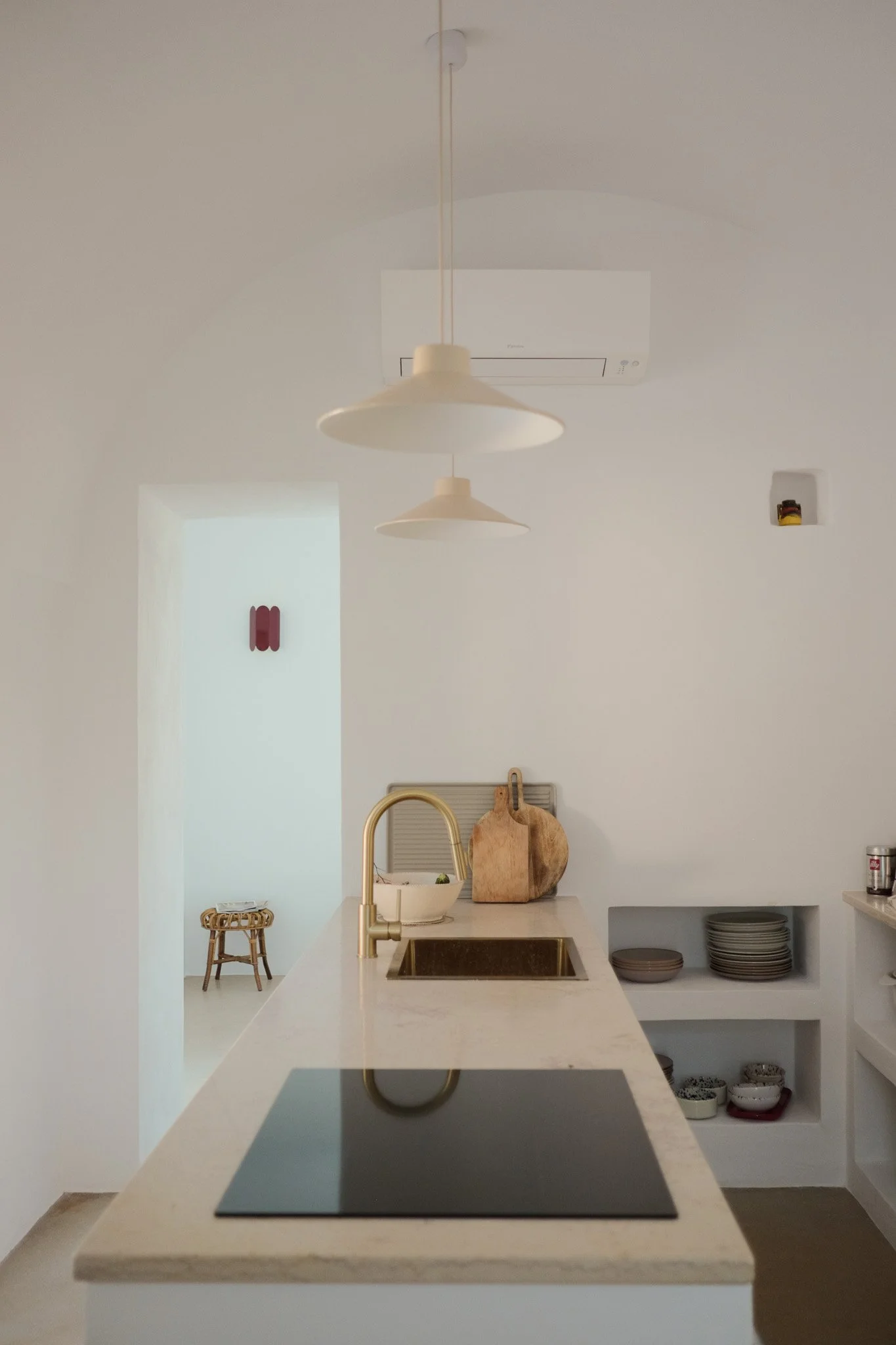 Bright kitchen with white walls and a marble countertop. A gold faucet over a built-in sink, stacks of bowls and plates on open shelves, and decorative items including a wooden cutting board and a bowl with green herbs. Hanging beige pendant lights a
