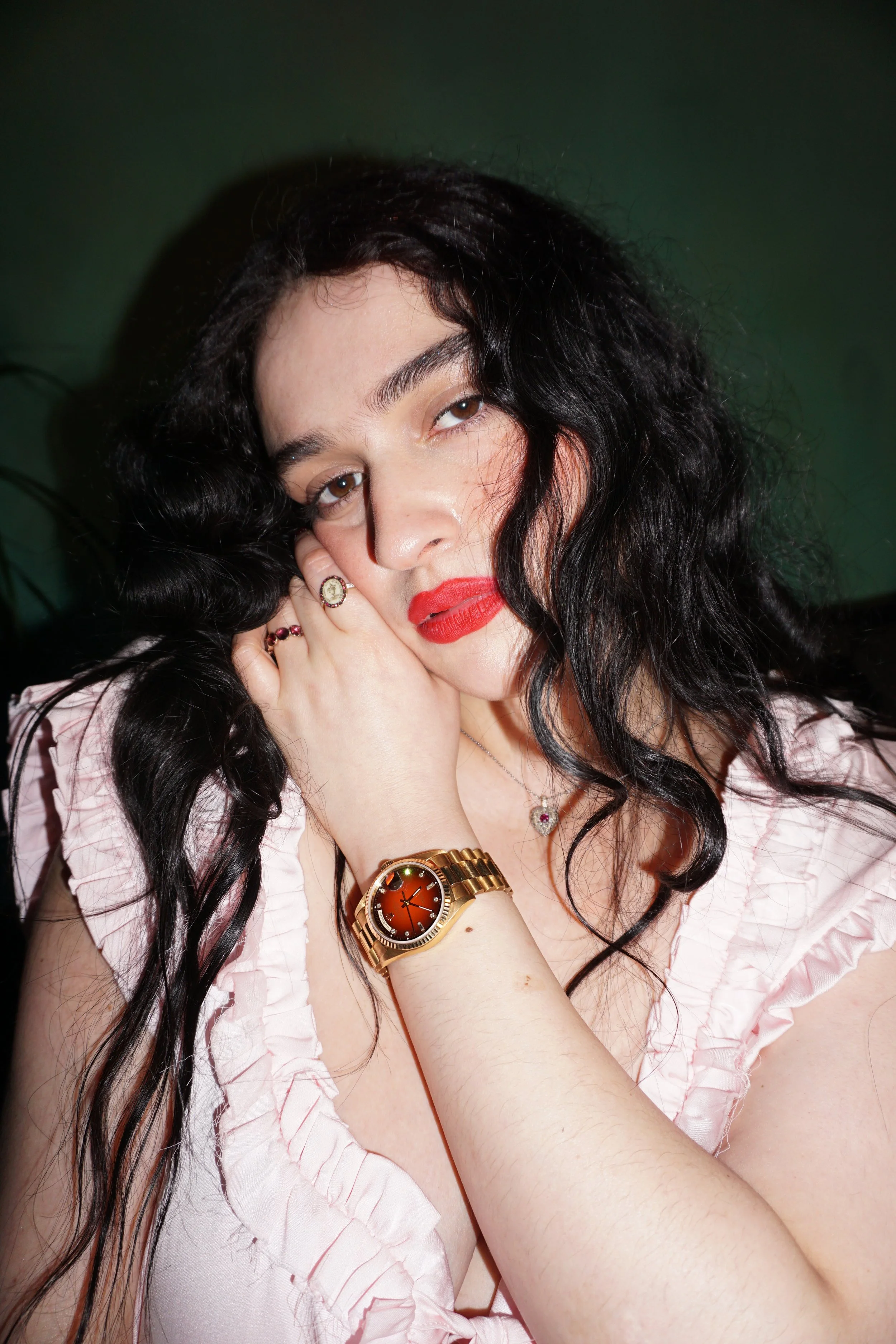 Close-up of a woman with long dark curly hair, wearing red lipstick, a pink satin ruffled blouse, a gold watch, a necklace, and rings, with her hand resting near her face against a dark green background.