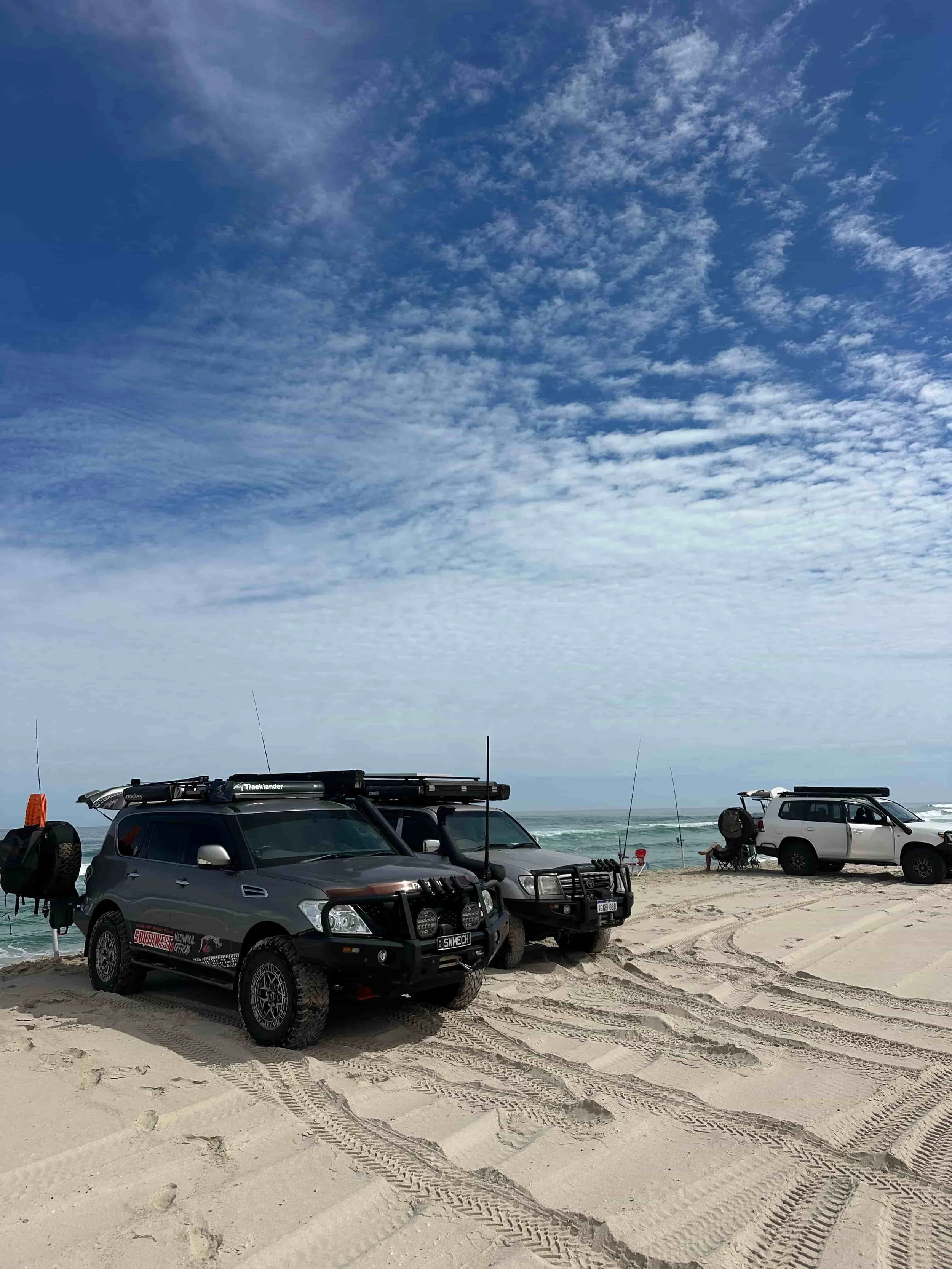Beach scene with four parked 4Wds on sandy shore, some with roof racks and off-road gear, beach chairs, and people fishing by the ocean. The sky is partly cloudy with a blue and white cloud pattern.