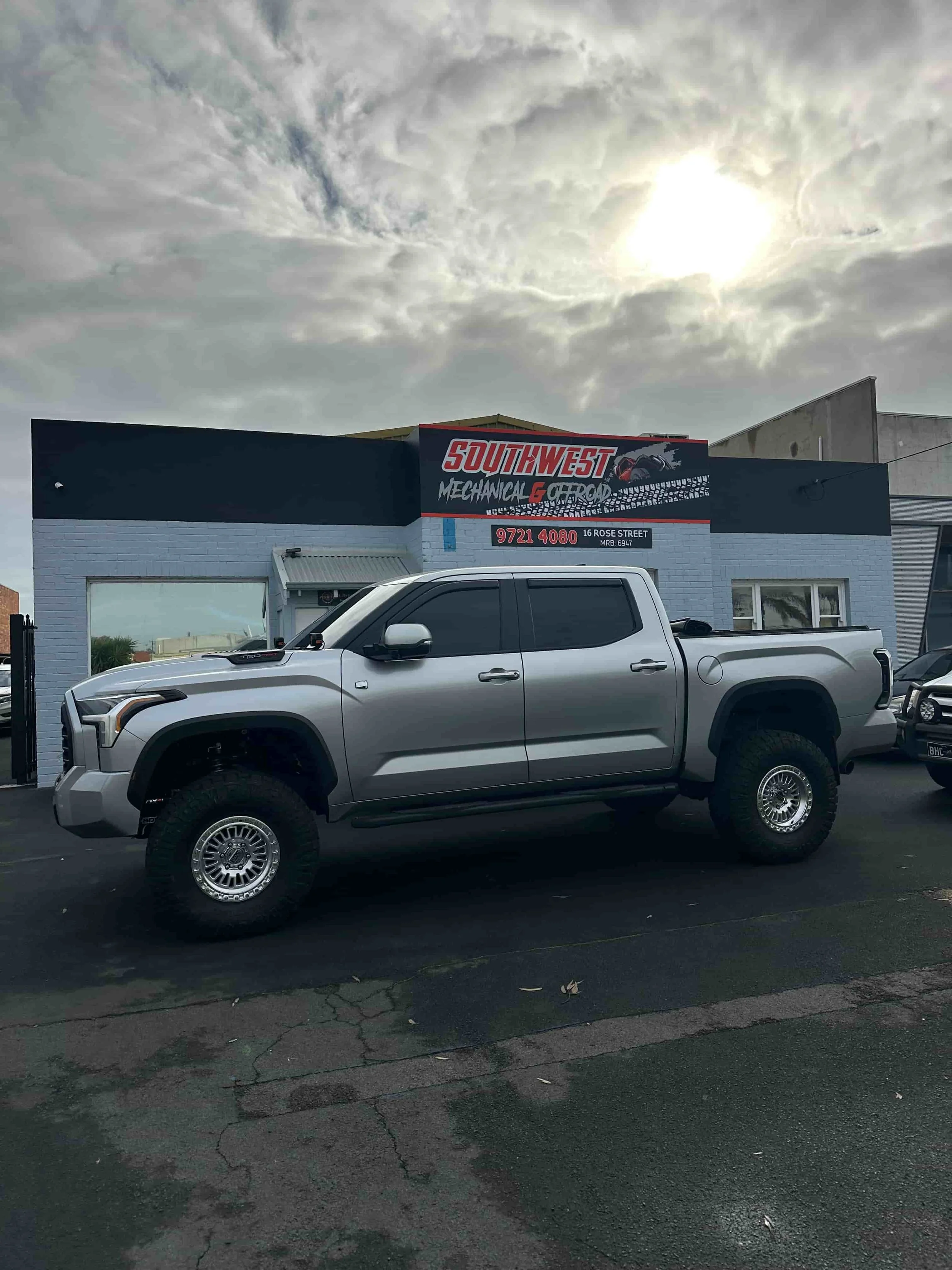 A silver 4WD parked in front of a building with a sign that reads 'Southwest Mechanical & Road'. The sky is cloudy with the sun partially obscured.