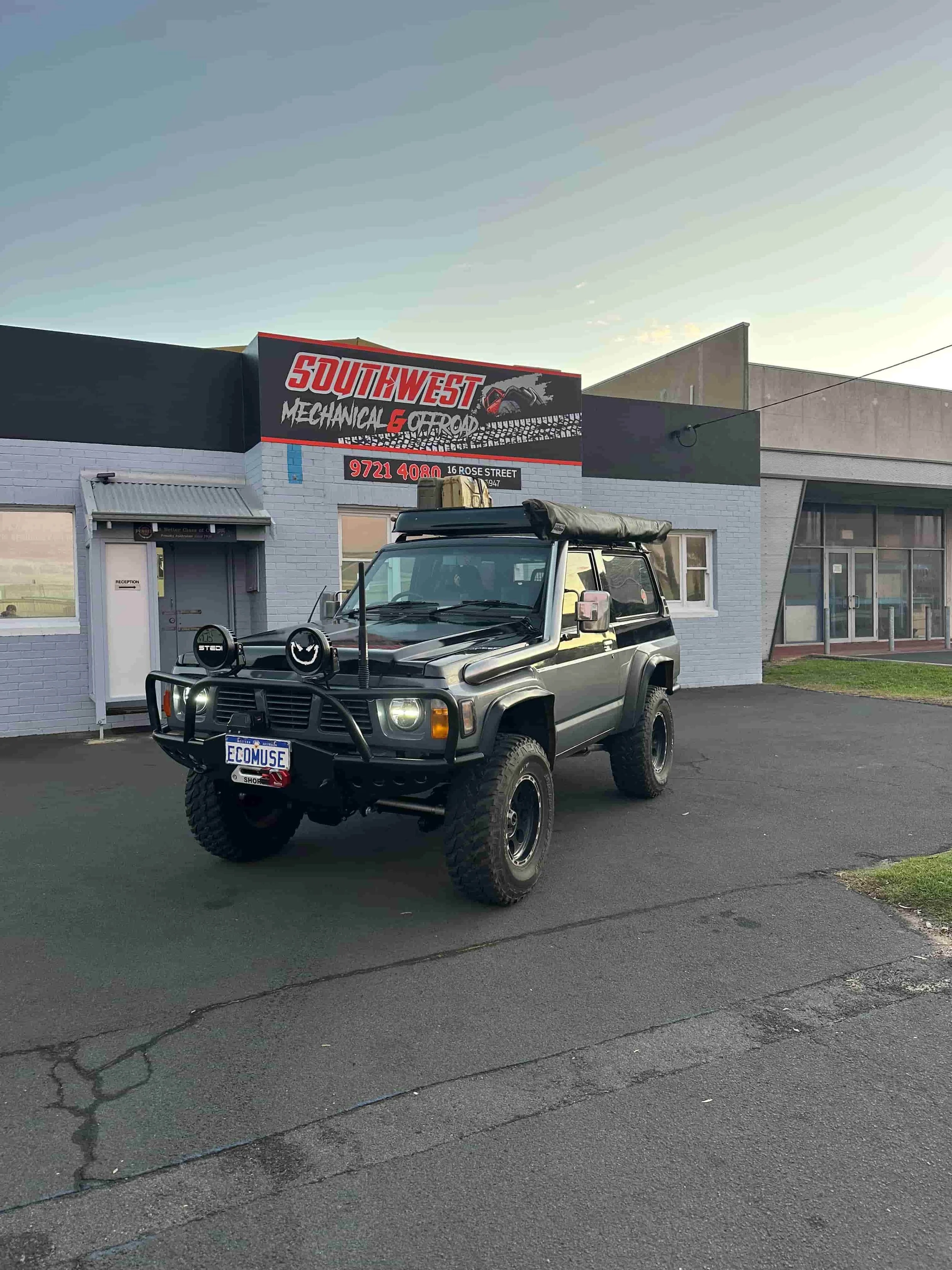 A black off-road vehicle with a roof rack, front bumper guard, and large tires parked in front of a grey building with a sign that reads 'Southwest Mechanical & Chrome'. The sign has red and black text and an image of a tire tread and an exhaust pipe, with a phone number and address below. The sky is clear, and the scene is lit by natural light, suggesting it is late afternoon or early evening.
