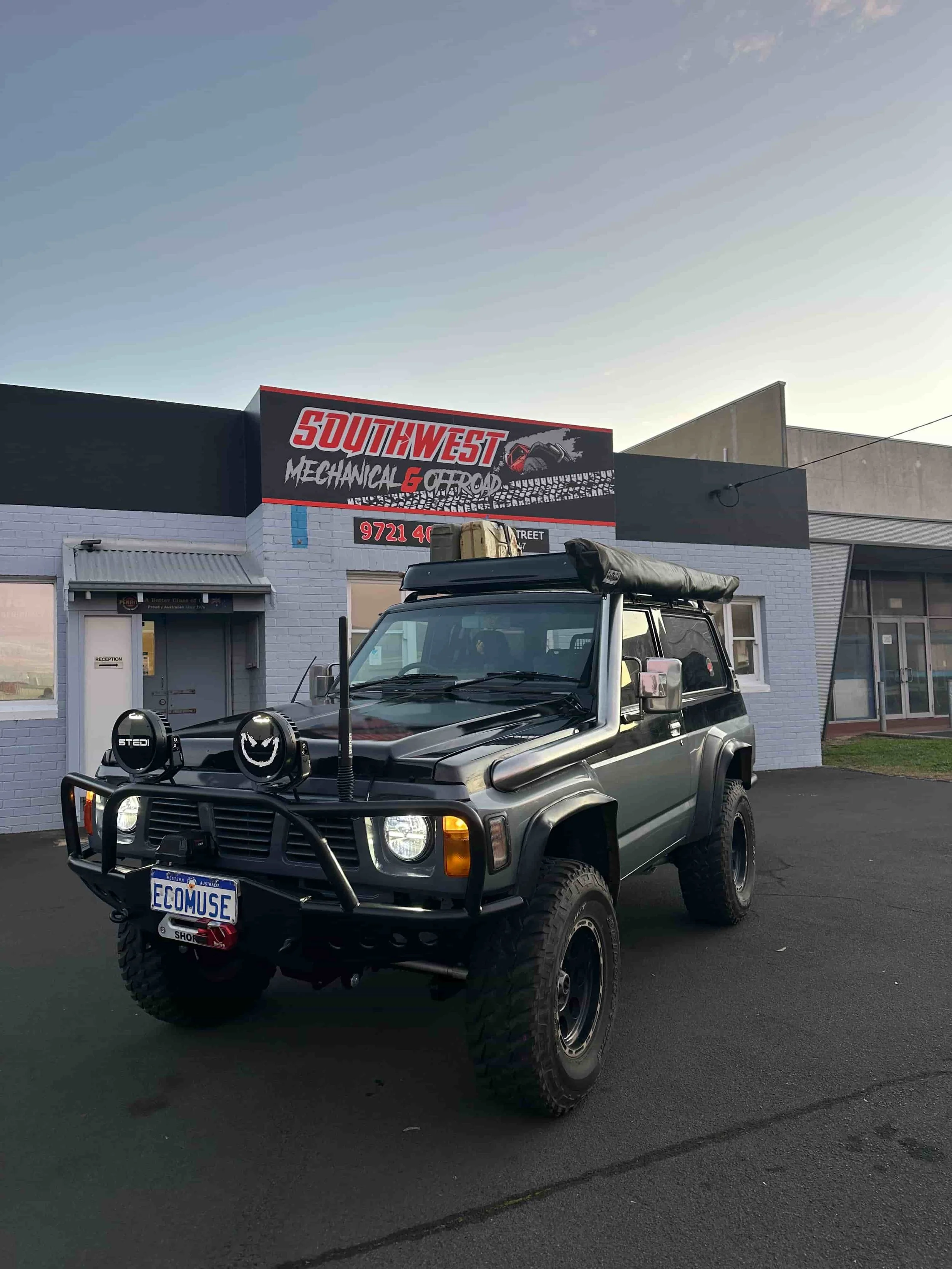 A black off-road vehicle with large tires, accessories, and a custom front bumper parked in front of a gray industrial building with a sign that reads 'Southwest Mechanical & Offroad.'