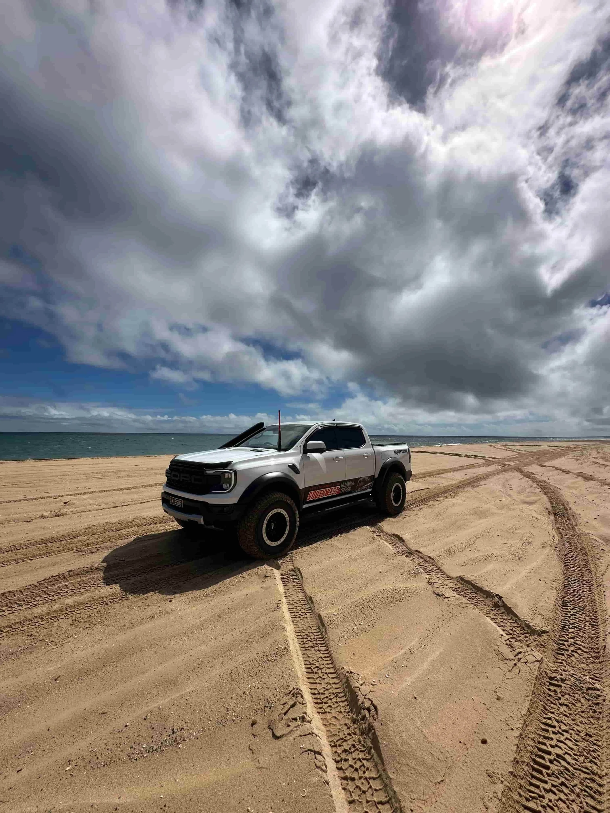 A white Ford Raptor 4WD parked on sandy terrain near a body of water under a cloudy sky.