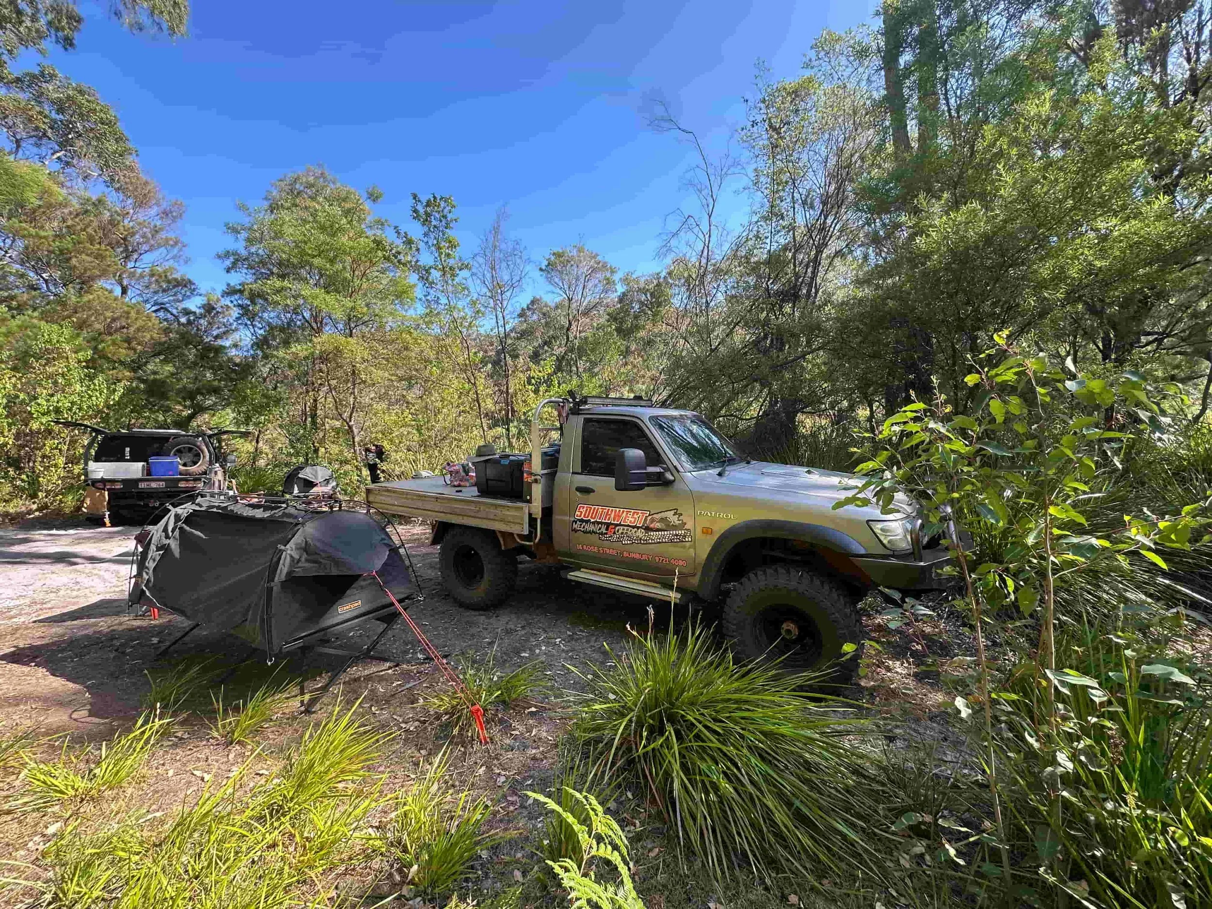 A campsite in a forest with trees and bushes, with off-road vehicles and camping gear including tents and a trailer.