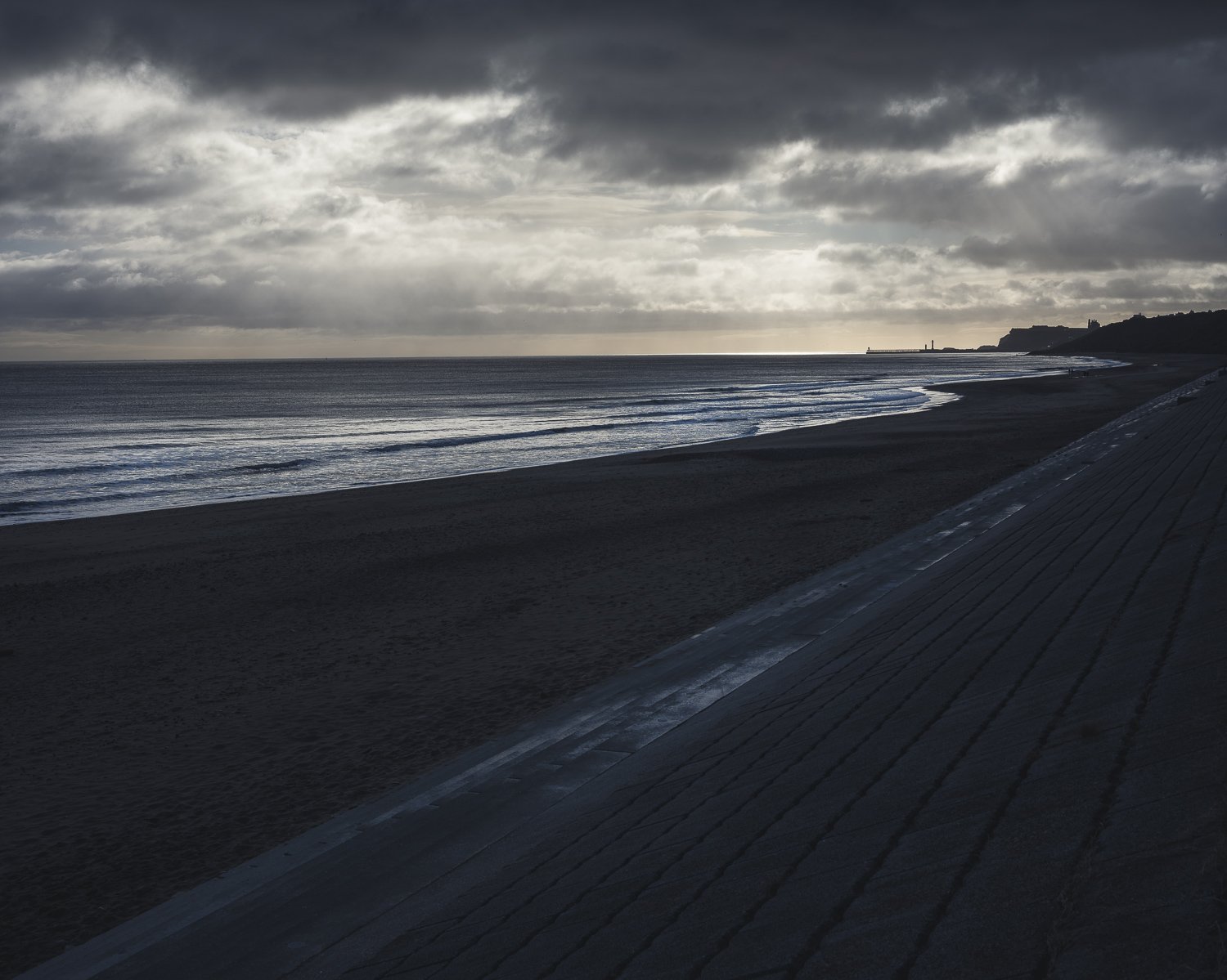 A cloudy beach scene with dark clouds overhead, calm waves on the shoreline, and a distant lighthouse on a headland.