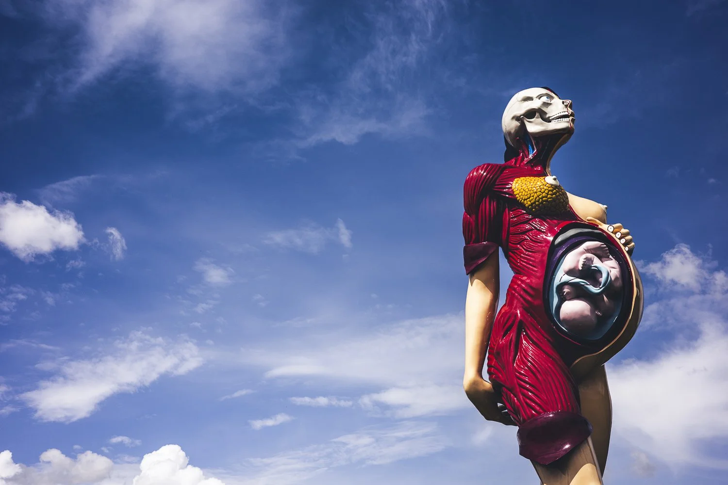 A large surreal sculpture of a woman with an open stomach exposing a fetus in the womb, with some anatomical details and exaggerated features, set against a blue sky with scattered clouds.