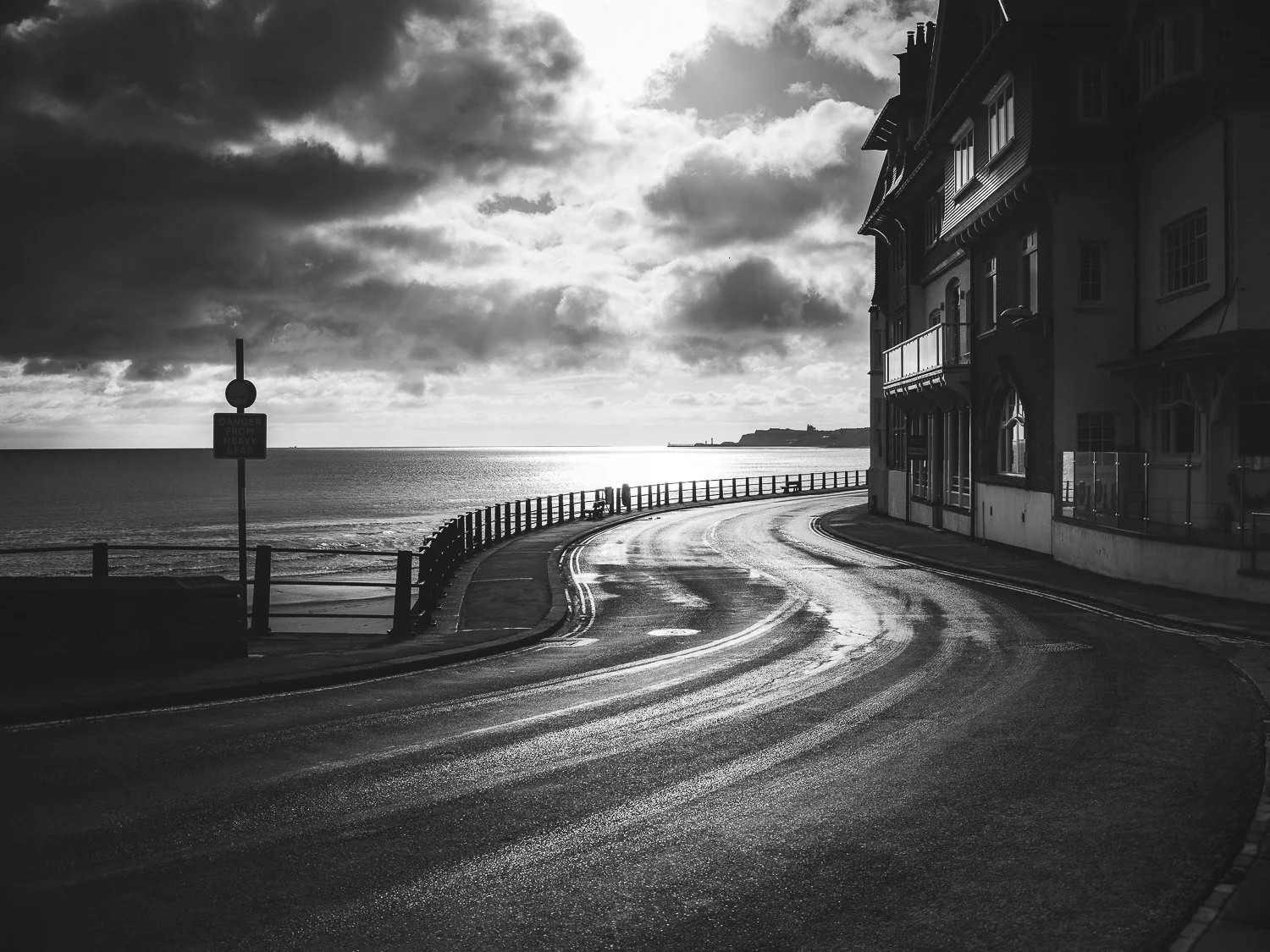 A black and white photo of a coastal road curving along the shoreline with a building on the right and an ocean view on the left. Dark clouds cover the sky, and sunlight reflects on the wet road surface.