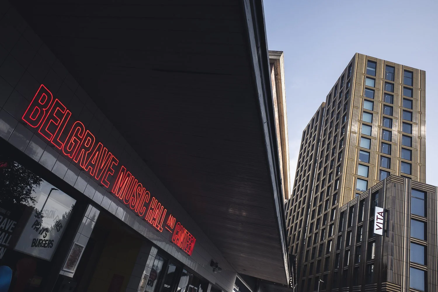 City street scene with a modern building, a neon sign reading "Belgraine Music Hall and Restaurant," and a tall office building in the background.