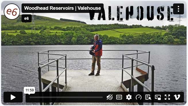 A man standing on a dock at Woodhead Reservoir, holding a camera, with green hills and trees in the background.