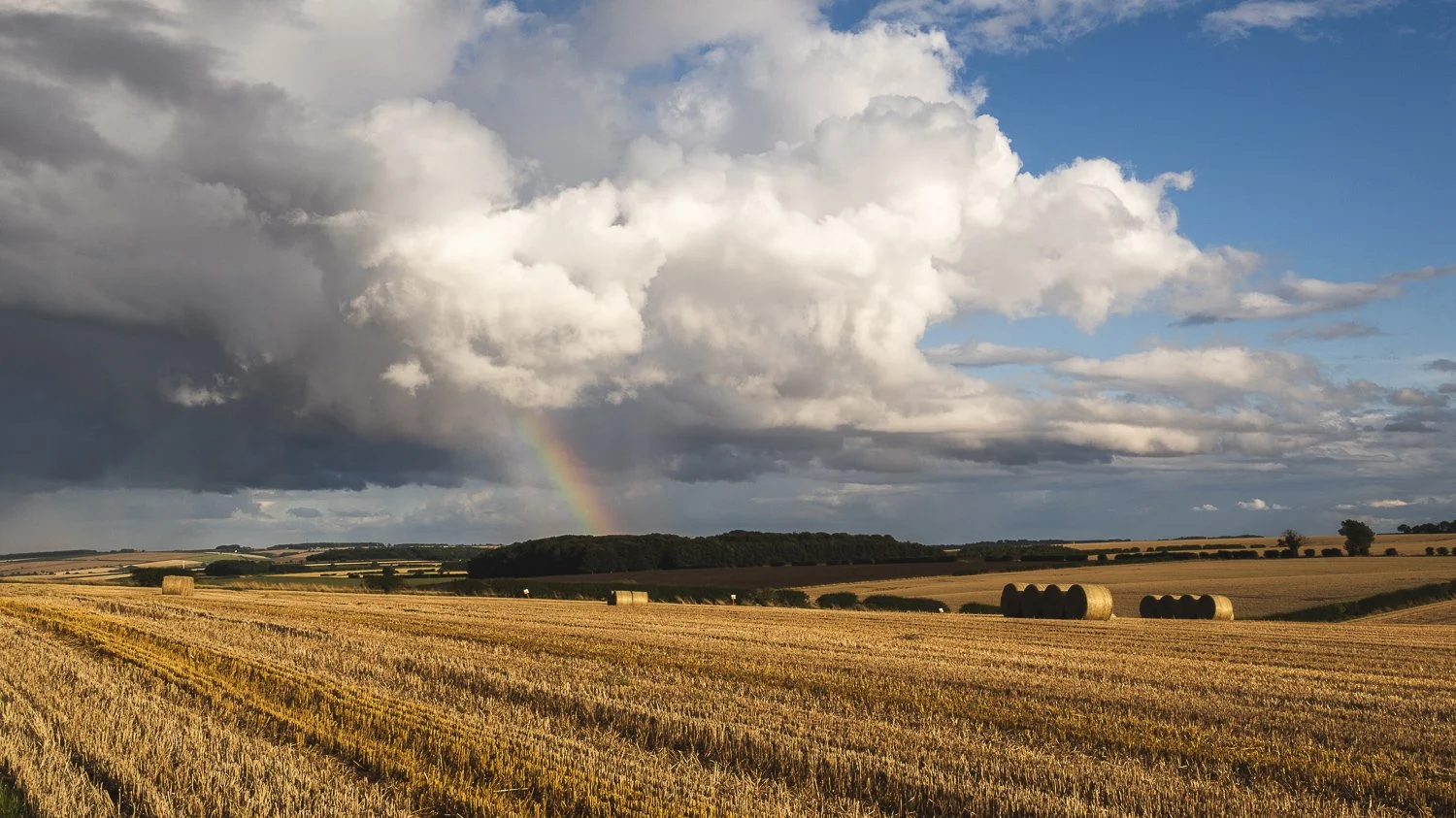 A vast farmland with hay bales scattered across golden fields, dark clouds in the sky with a rainbow in the distance.