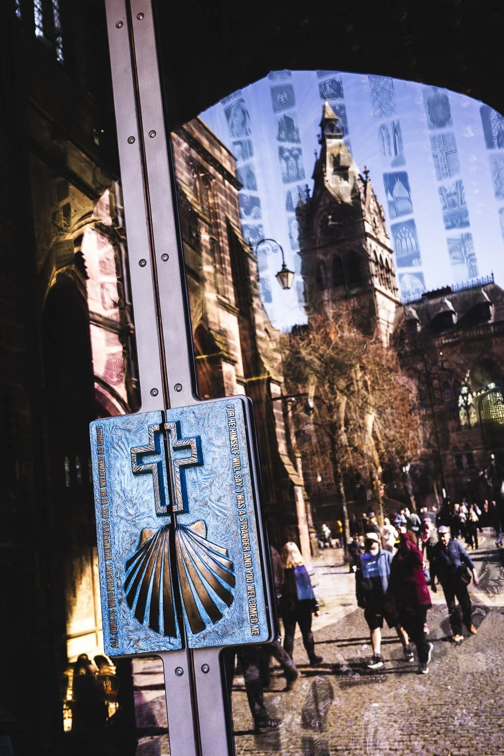 Reflection of a historic church tower and city street scene with people, seen through a glass with a religious sticker attached.