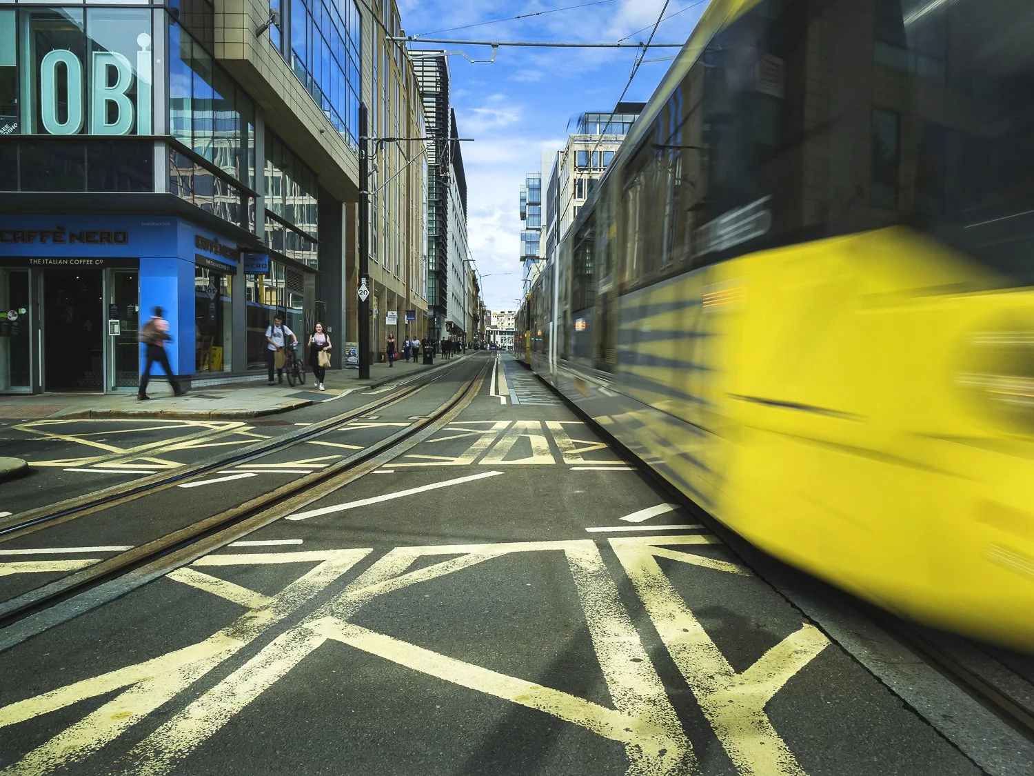 A city street scene with a yellow tram moving on tracks, pedestrians walking on the sidewalk, modern buildings with glass facades, and a blue sky with some clouds.